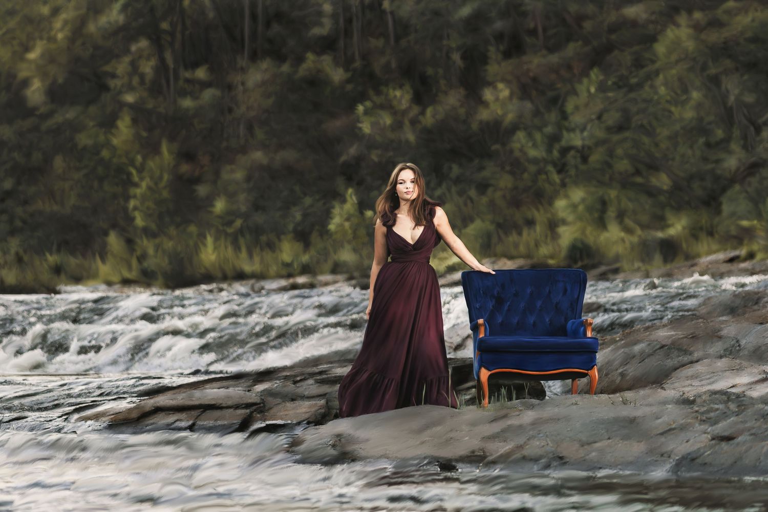 Woman in burgundy gown stands beside blue velvet chair on rocky riverside at sunset.