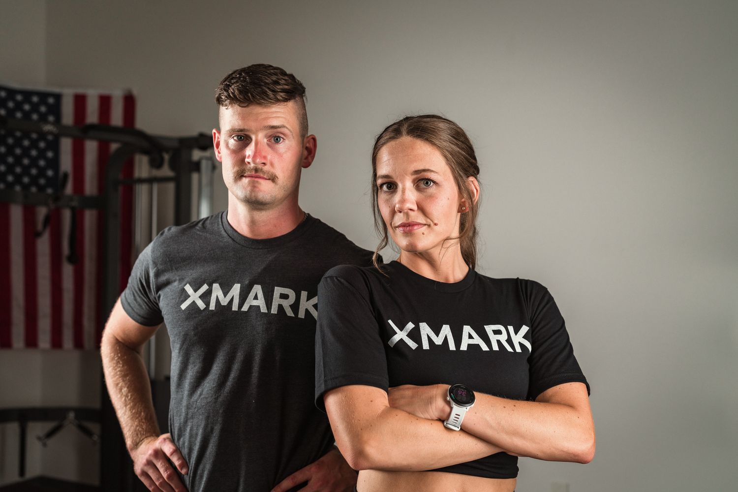 Man and woman in matching XMARK shirts posing seriously in a gym setting.