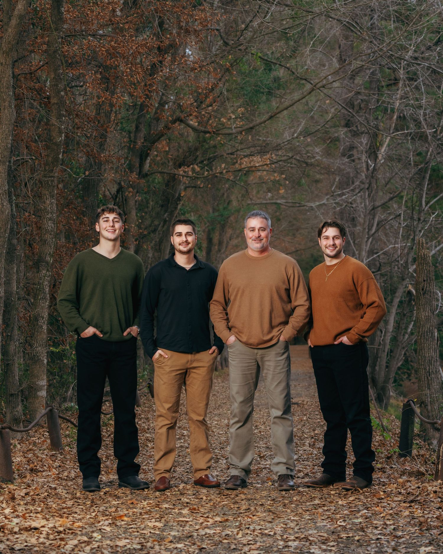 Four males stand together smiling on a leaf-covered path surrounded by autumn trees.