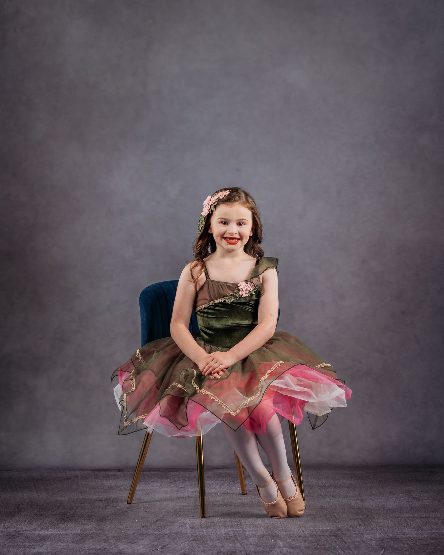 Smiling young girl in green and pink ballet costume sits on blue chair for dance portrait.