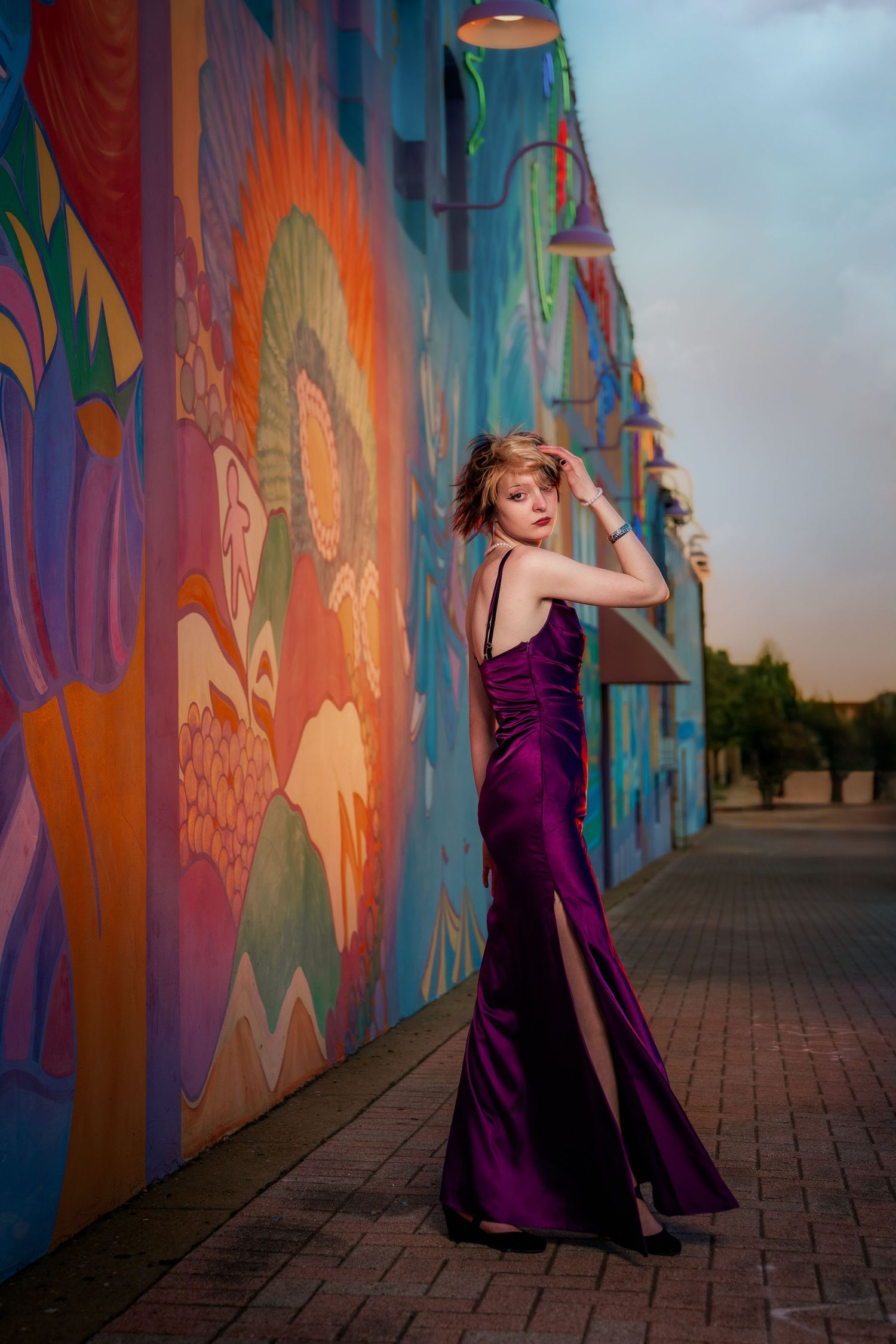 Woman in elegant purple satin gown poses beside vibrant colorful street mural at dusk.