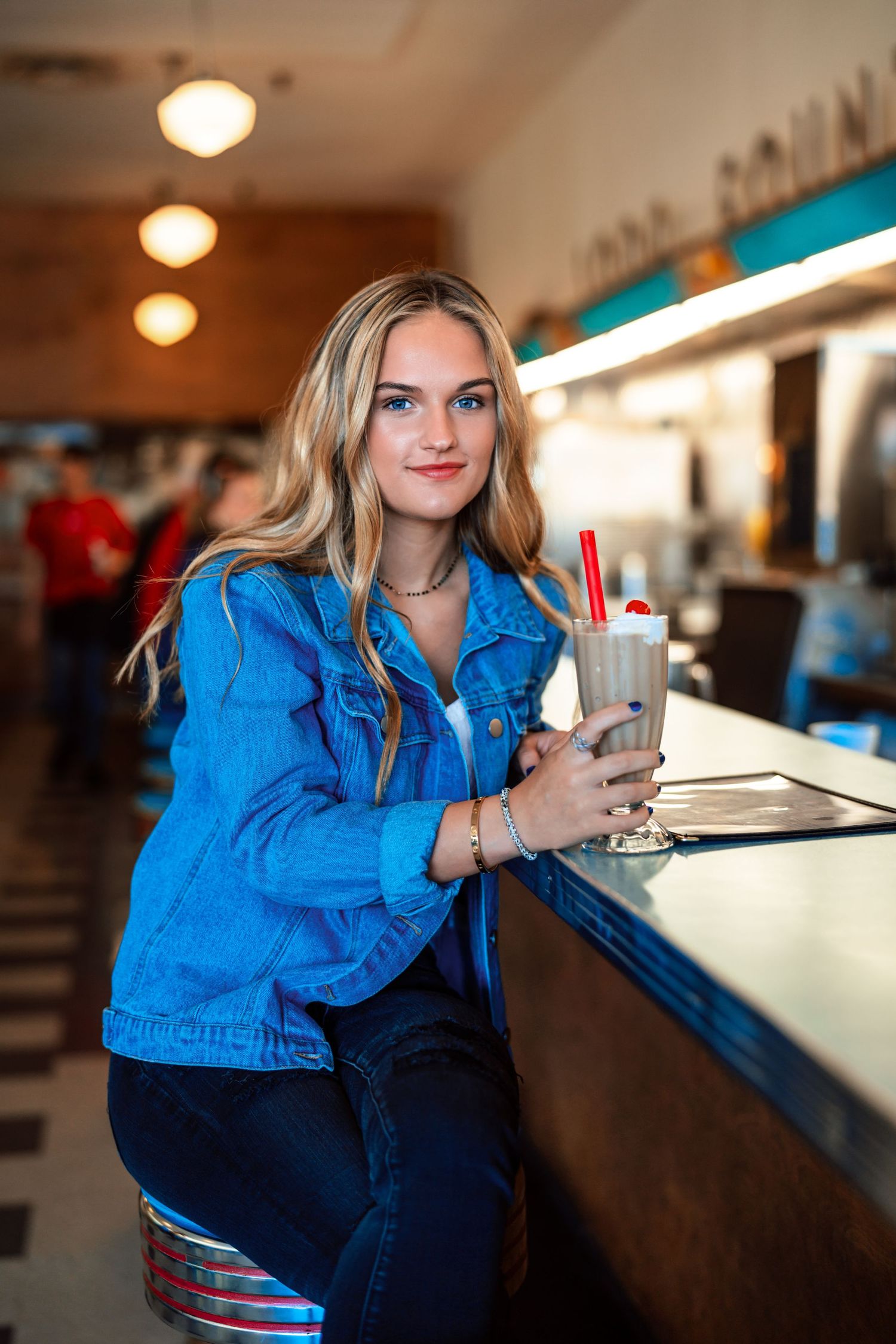 Blonde teen girl in a blue denim jacket holds a milkshake at a classic American diner counter.