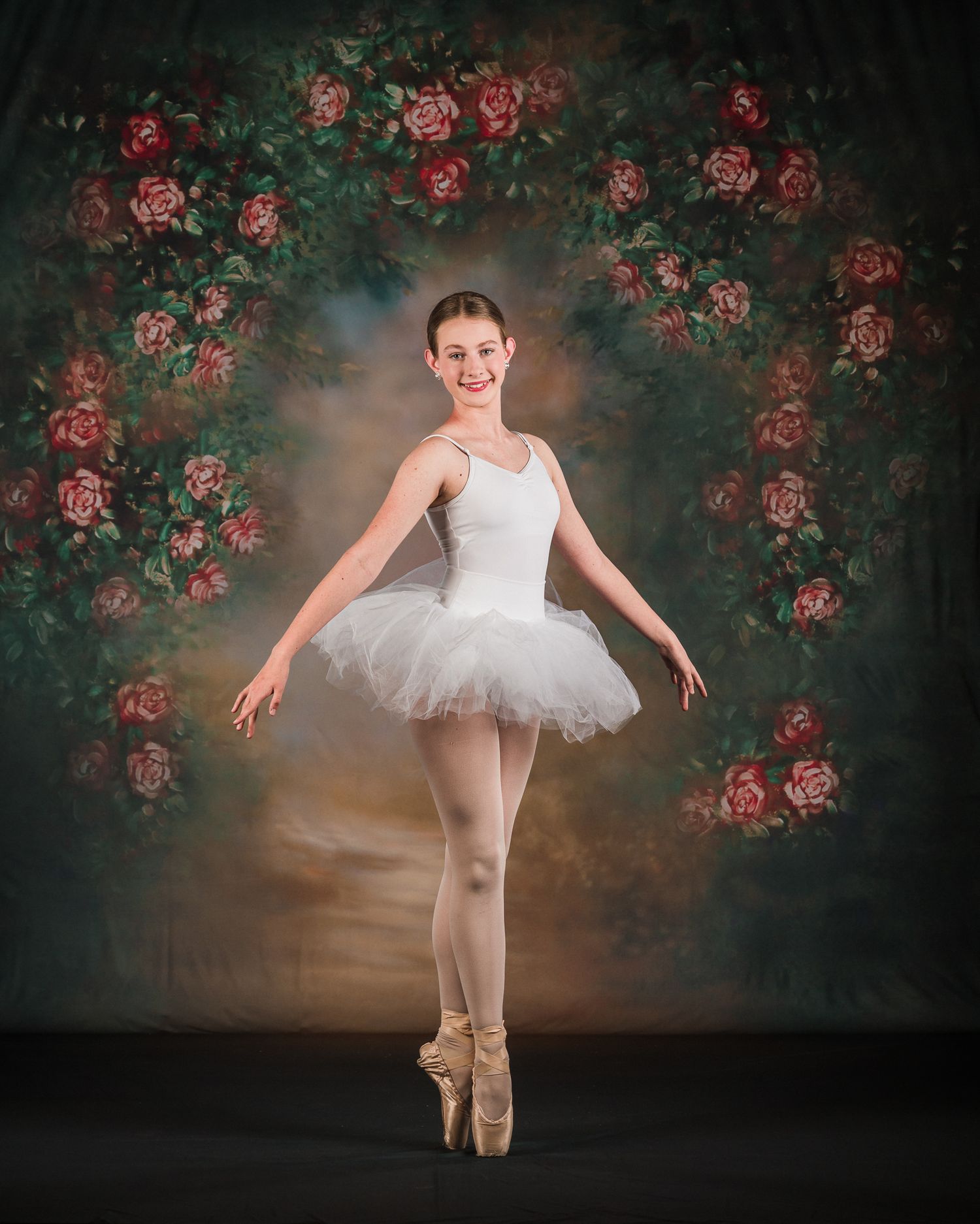 Young ballerina in white tutu on pointe smiling before romantic floral rose backdrop.
