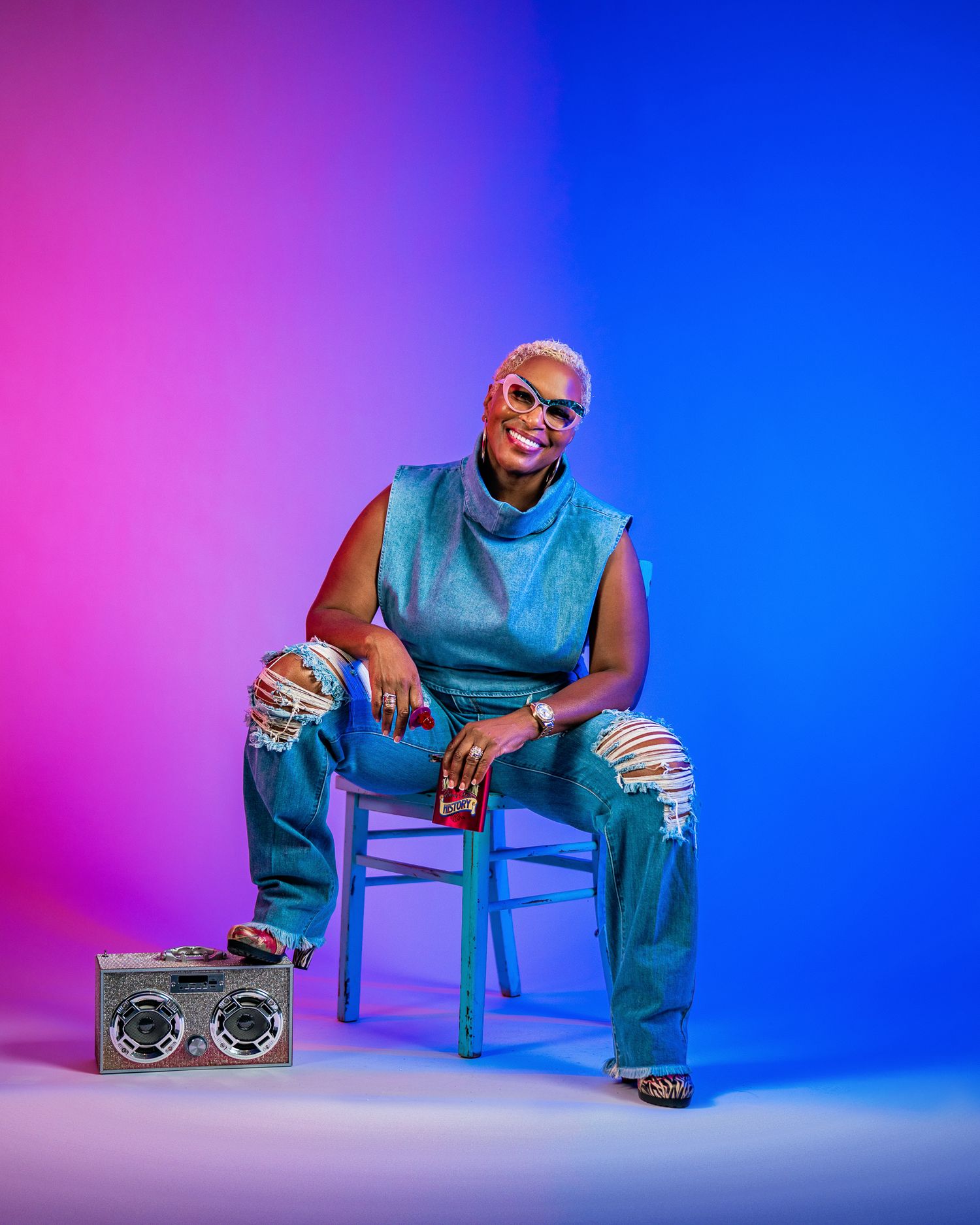 Confident woman in teal outfit sitting on chair with boombox on colorful background.