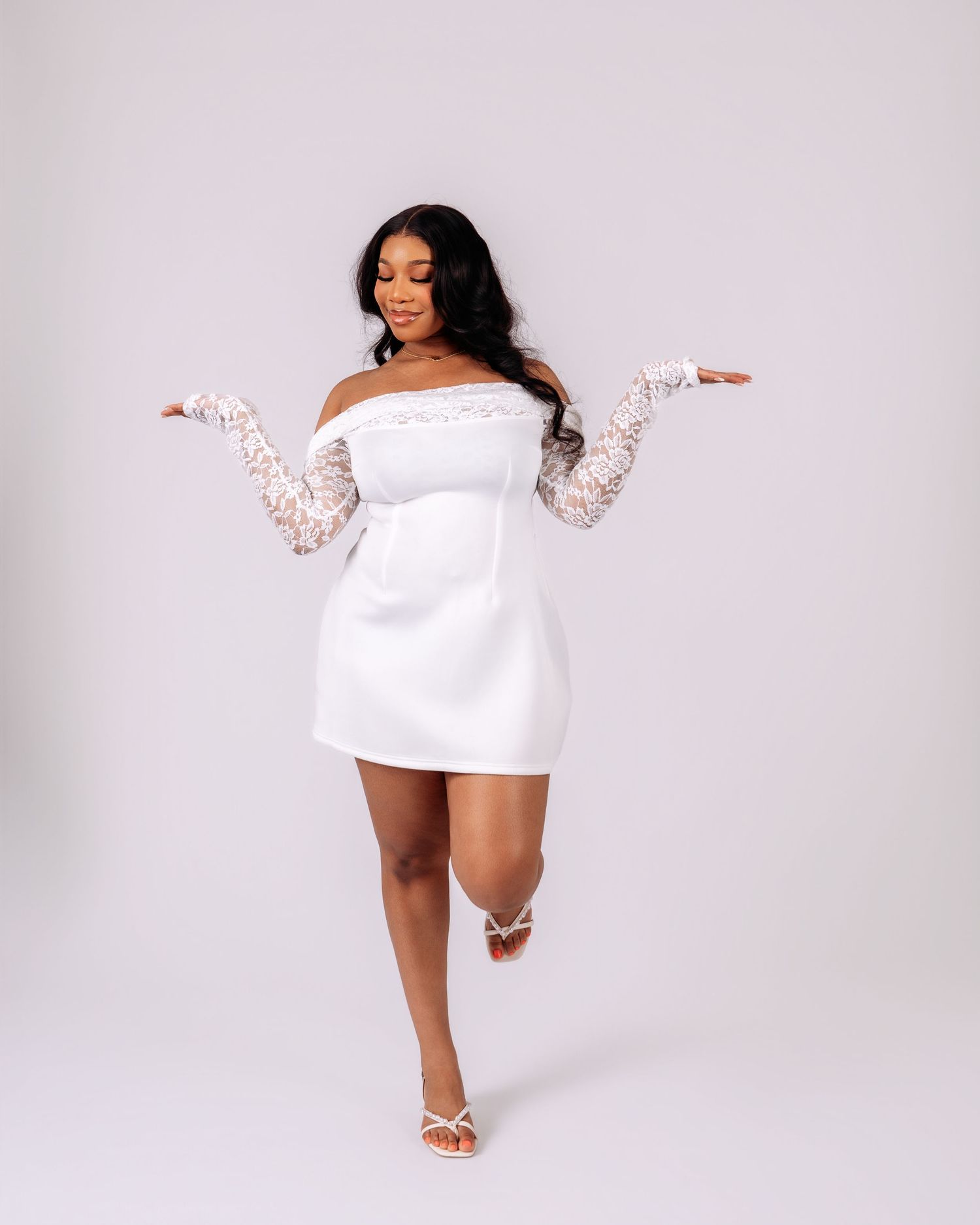 Woman in white off-shoulder mini dress with lace sleeves and strappy heels posing playfully on light background.