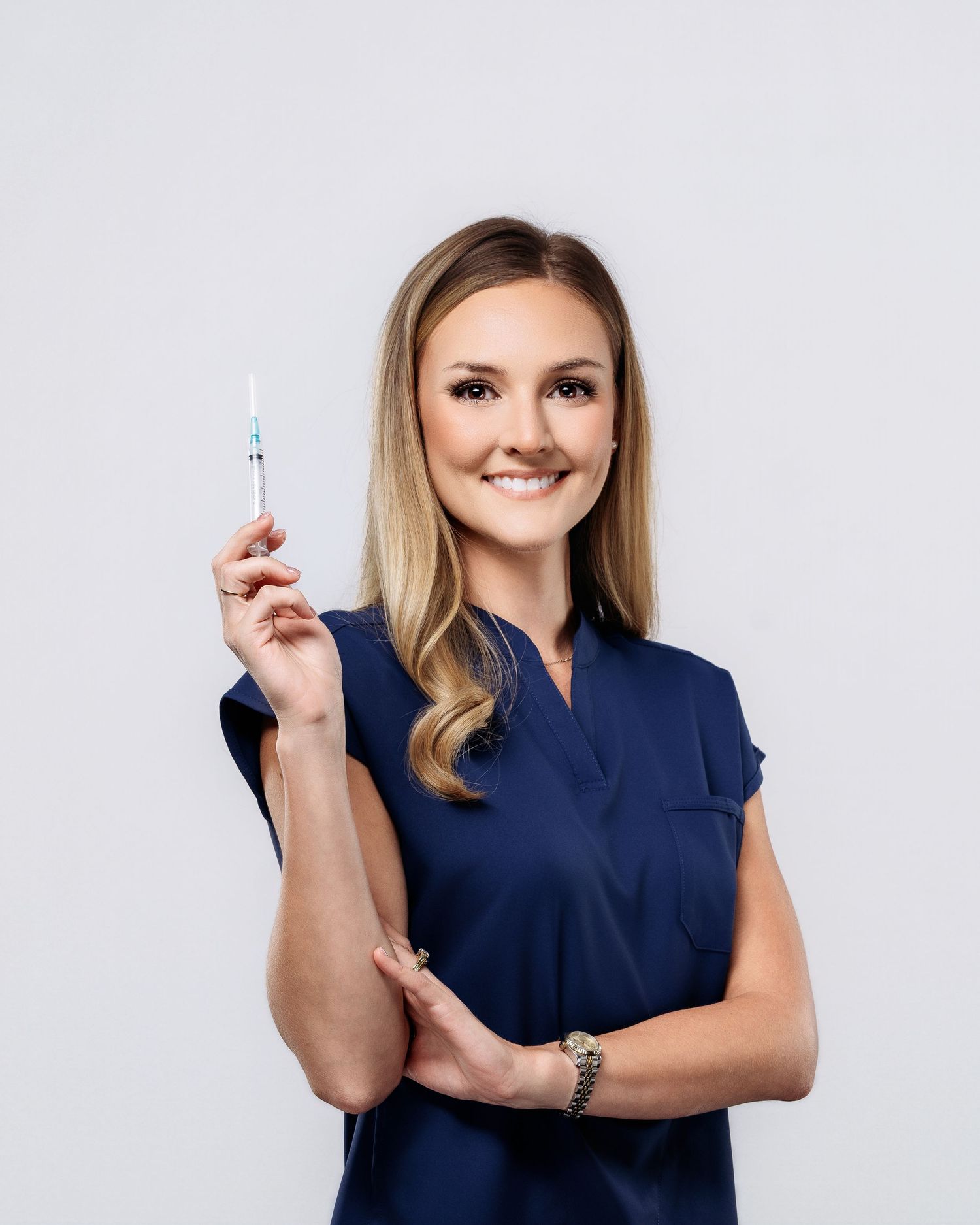 Smiling blonde woman in navy scrubs holding syringe in professional medical portrait.
