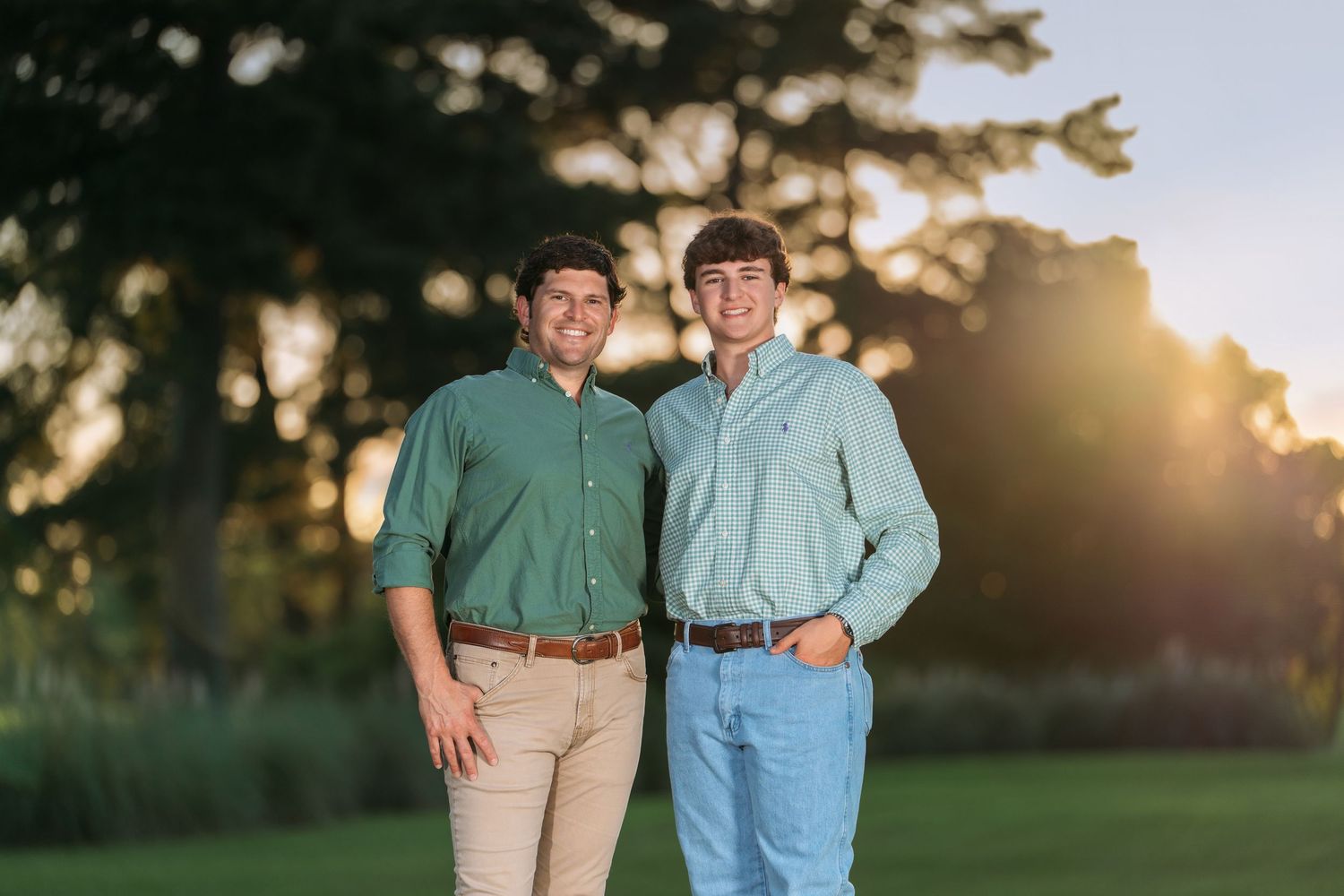 Two young men in matching green shirts smile together on a green lawn at golden hour.