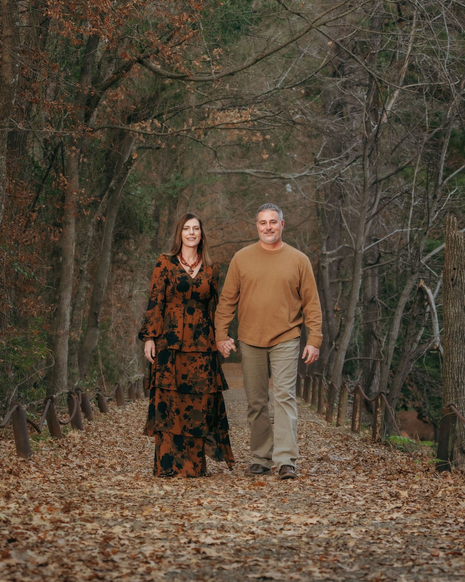 Smiling couple strolling hand-in-hand down a leaf-covered wooded path in warm autumn earth-tone outfits.
