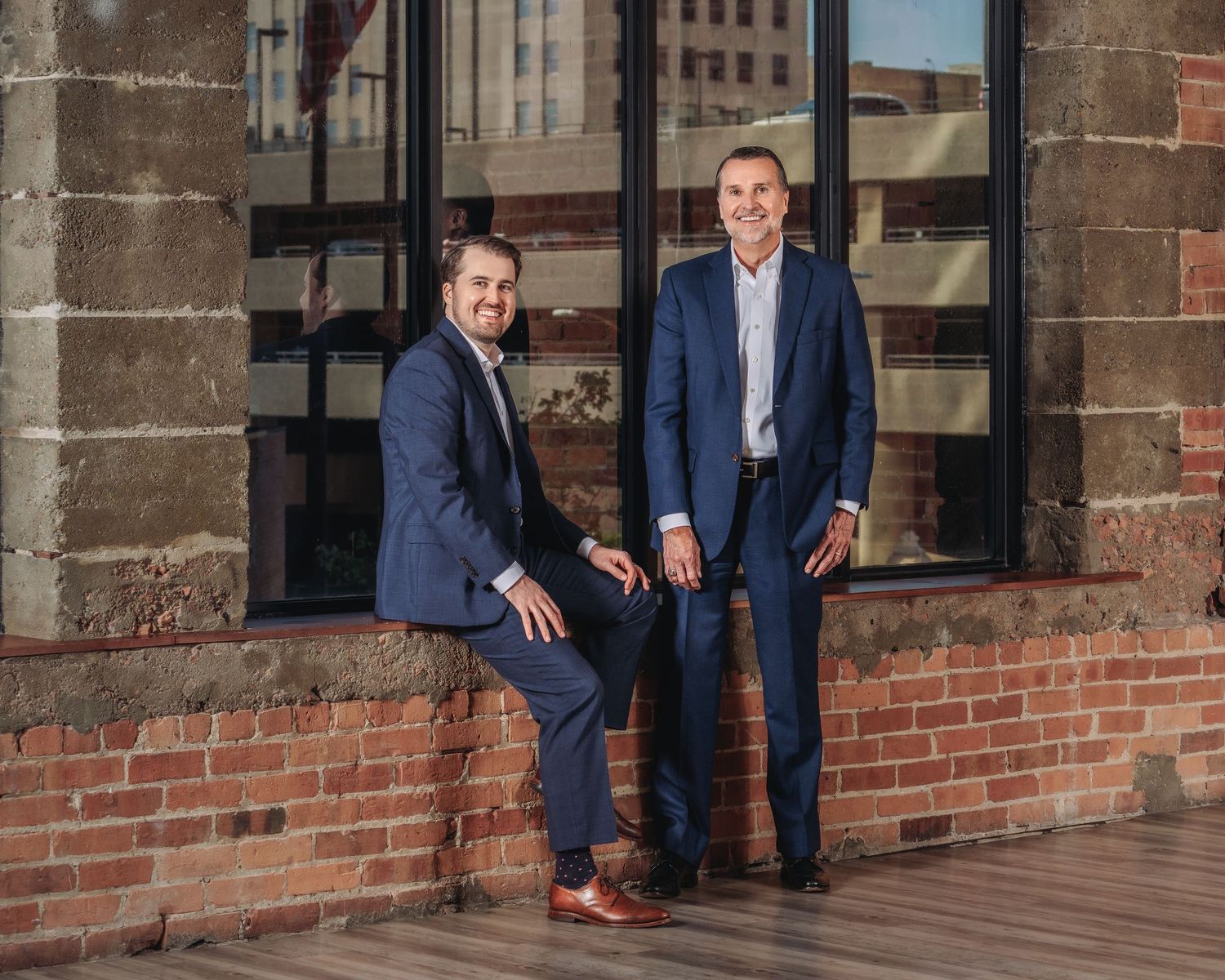 Two professional men in blue suits posing outdoors against a brick wall and large windows.