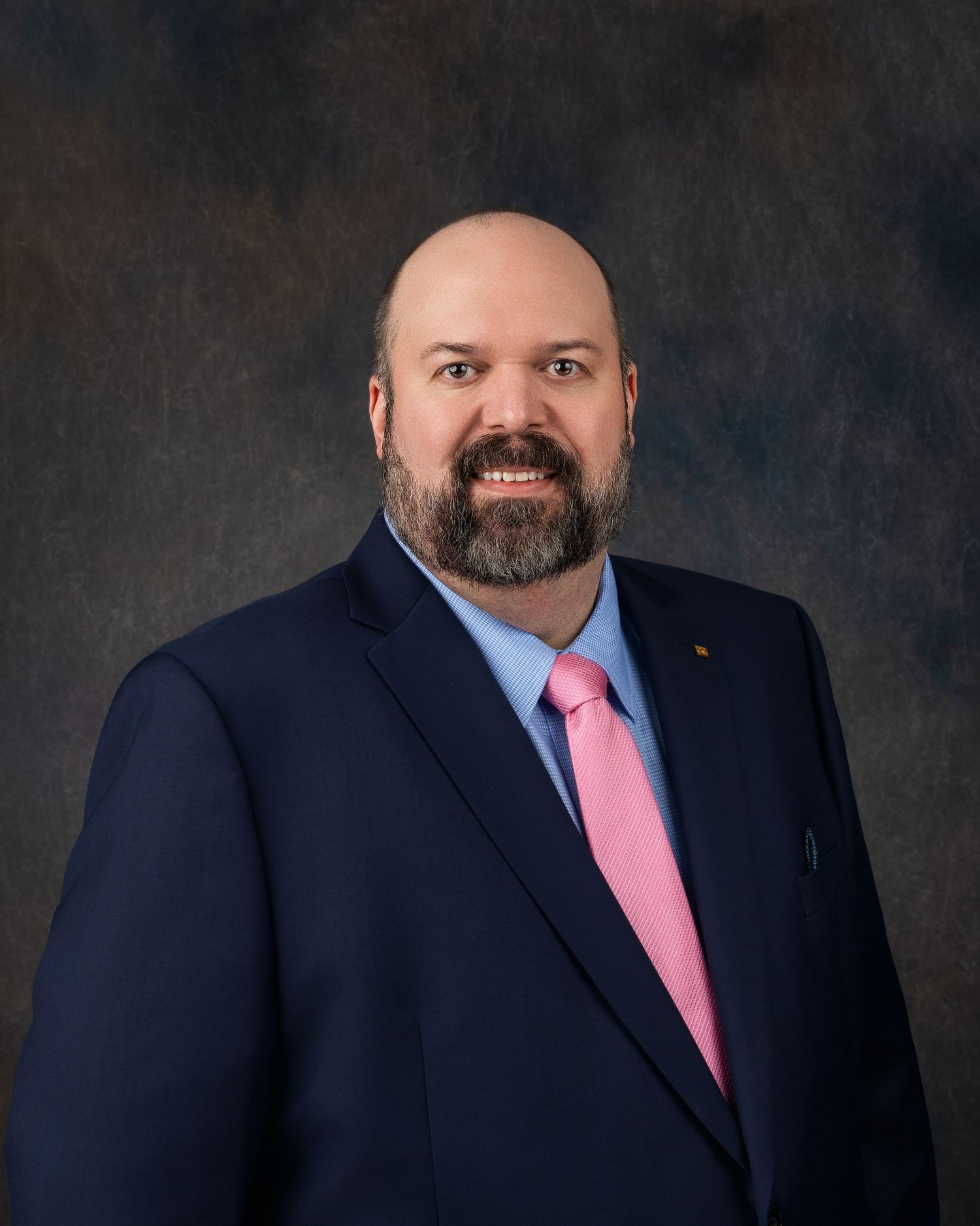 Professional headshot of a smiling bald man with a beard wearing a navy suit and pink tie.