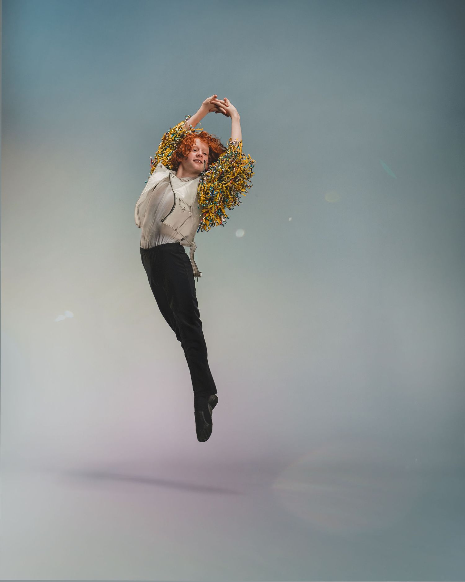 A red-haired dancer leaps gracefully mid-air, wearing black pants and a white top adorned with yellow flowers.