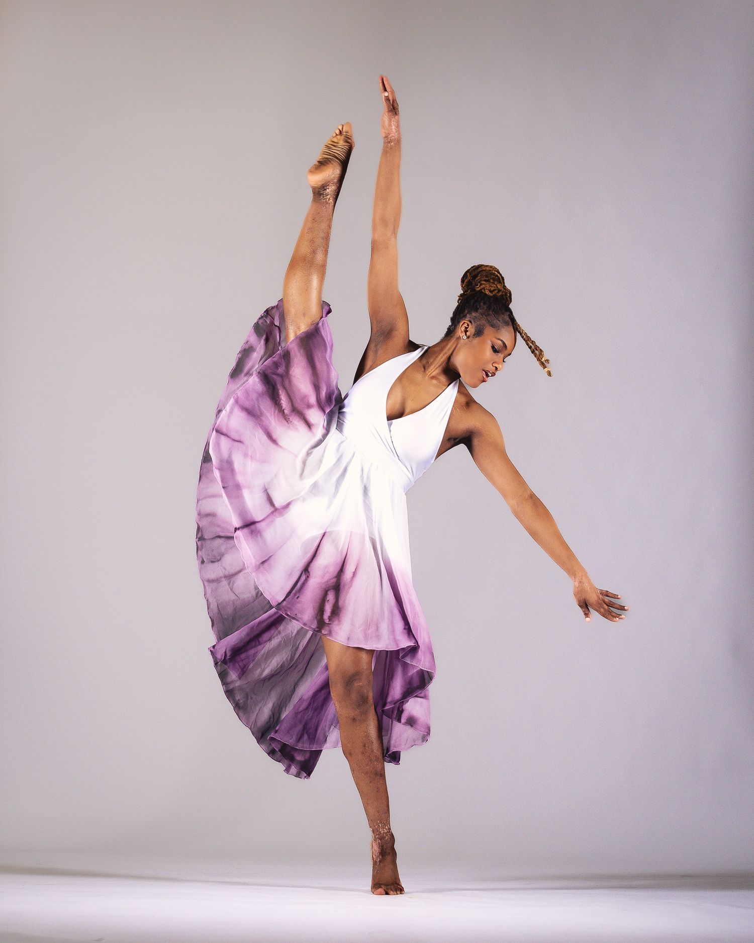 Contemporary dancer in purple tie-dye dress performing dramatic leg extension in studio.