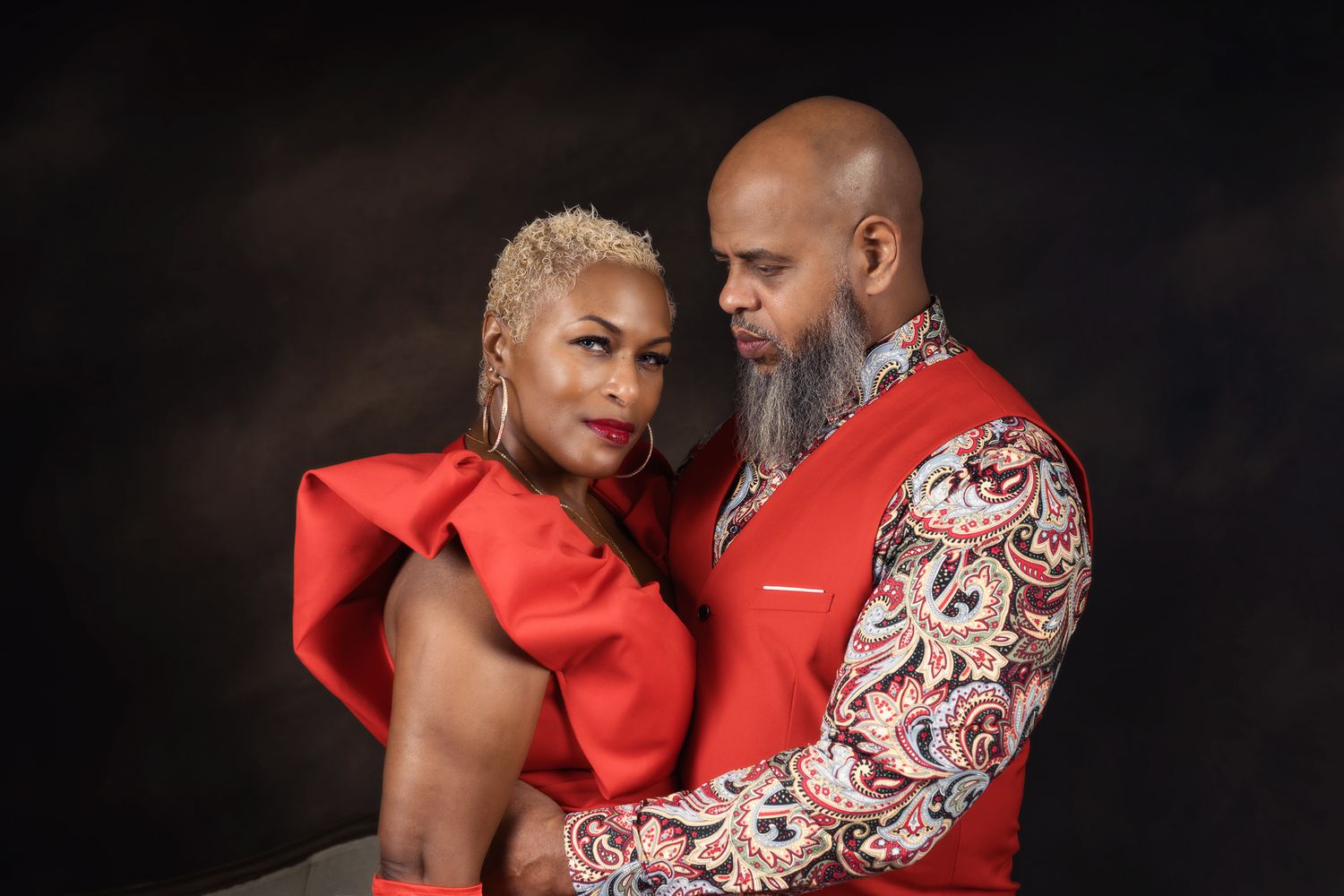 Stylish couple in red and patterned outfits posing together on dark background.
