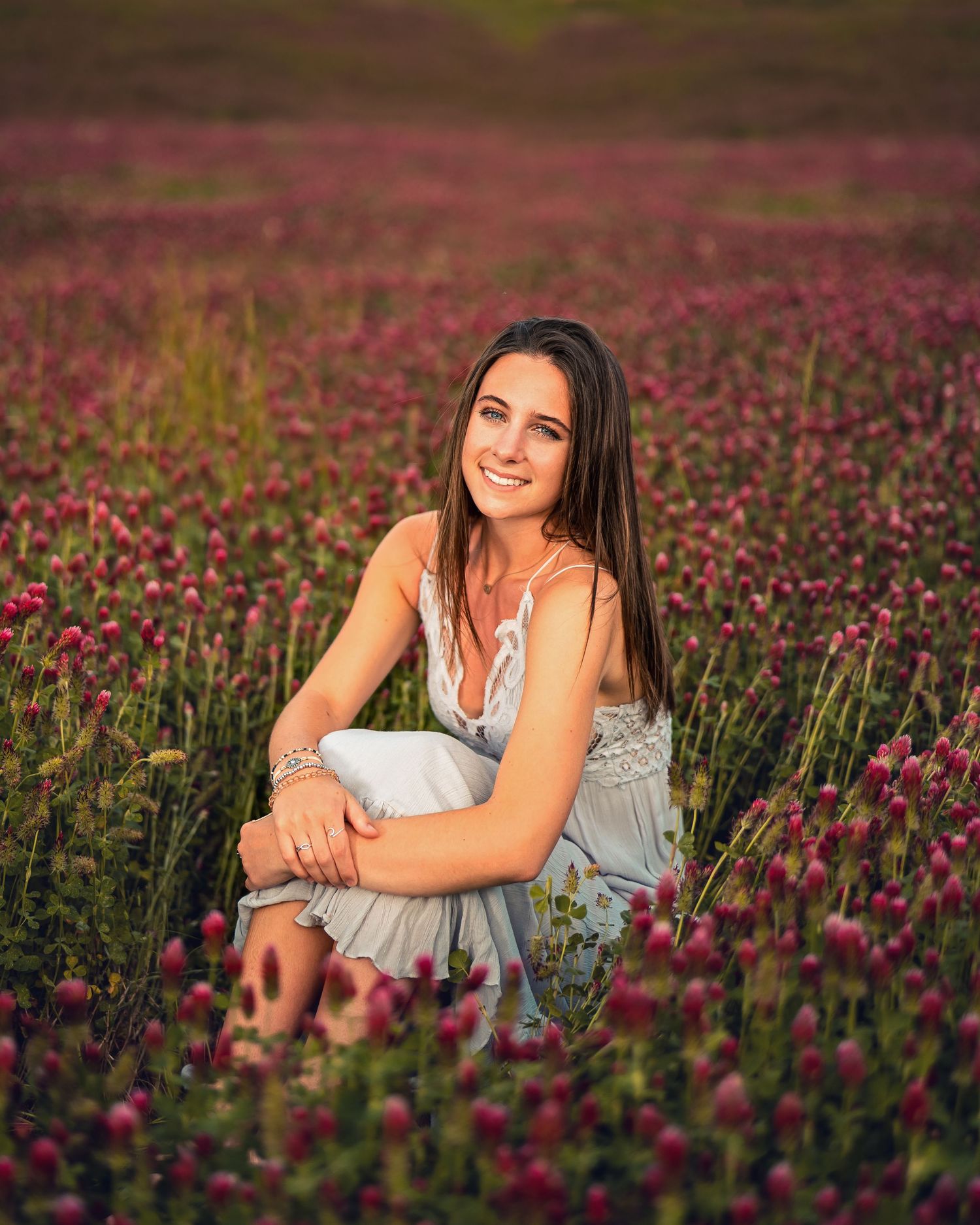 Smiling brunette woman in a light blue dress sits in a vibrant field of pink wildflowers.