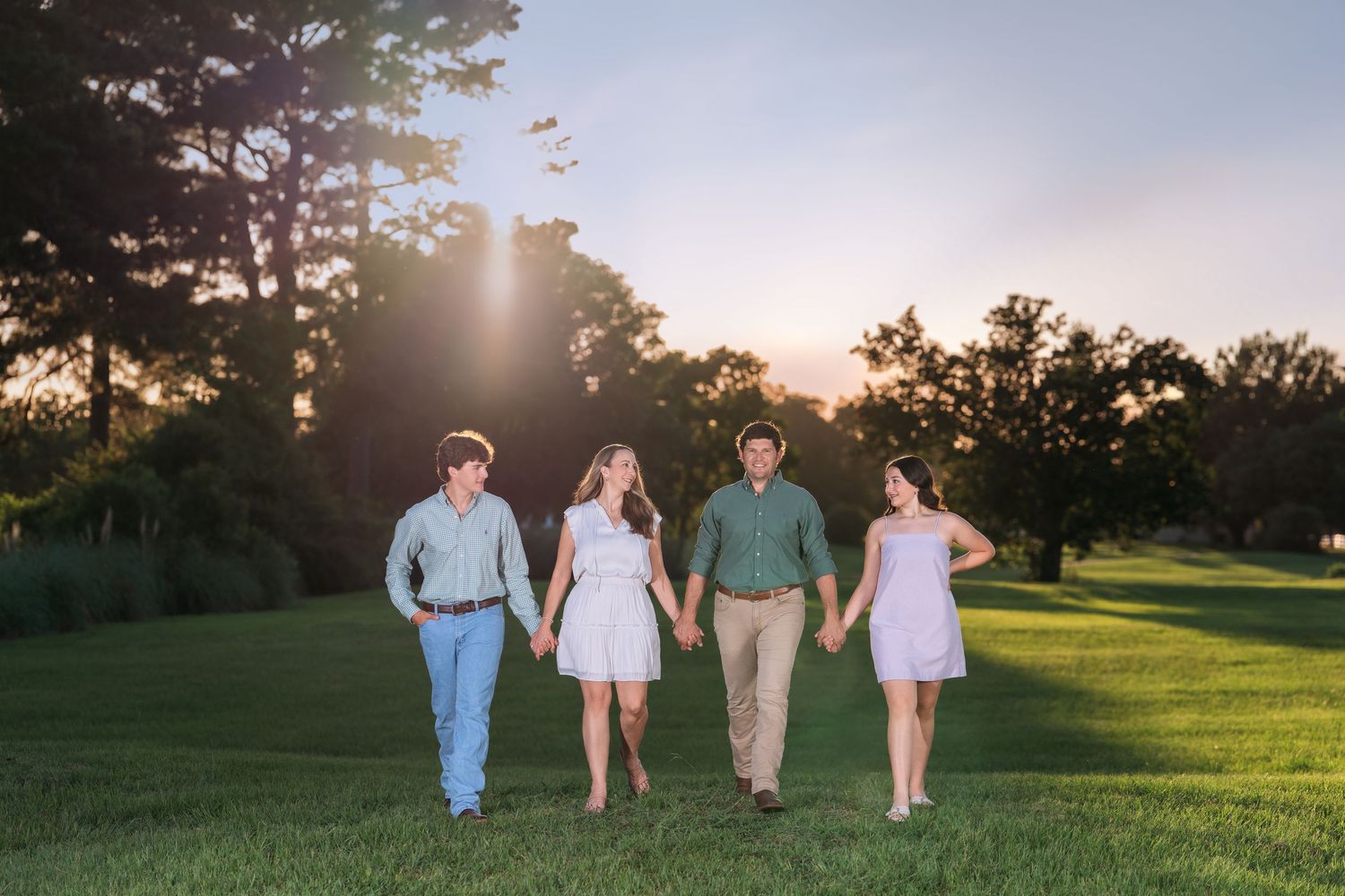 A group of four people hold hands and walk together across a sunlit green lawn at sunset.
