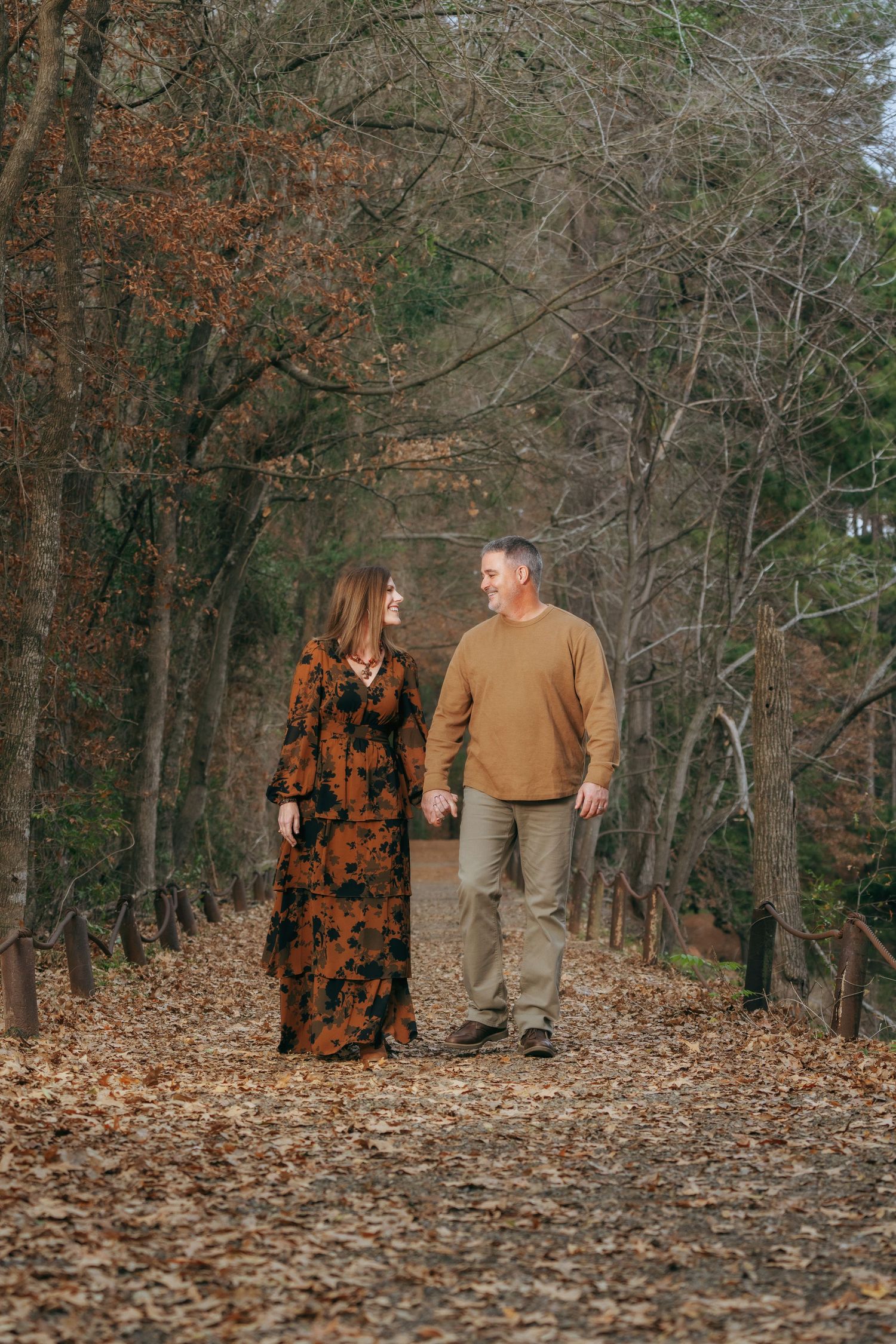 Couple walking hand-in-hand along a beautiful autumn forest path lined with bare trees and fallen leaves.