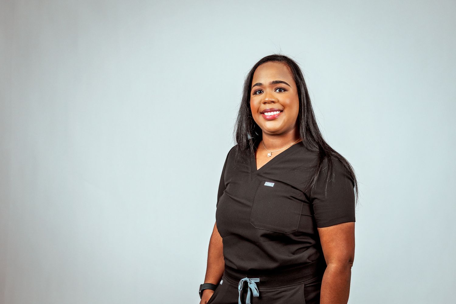 Healthcare professional in black scrubs smiling confidently against light gray background.