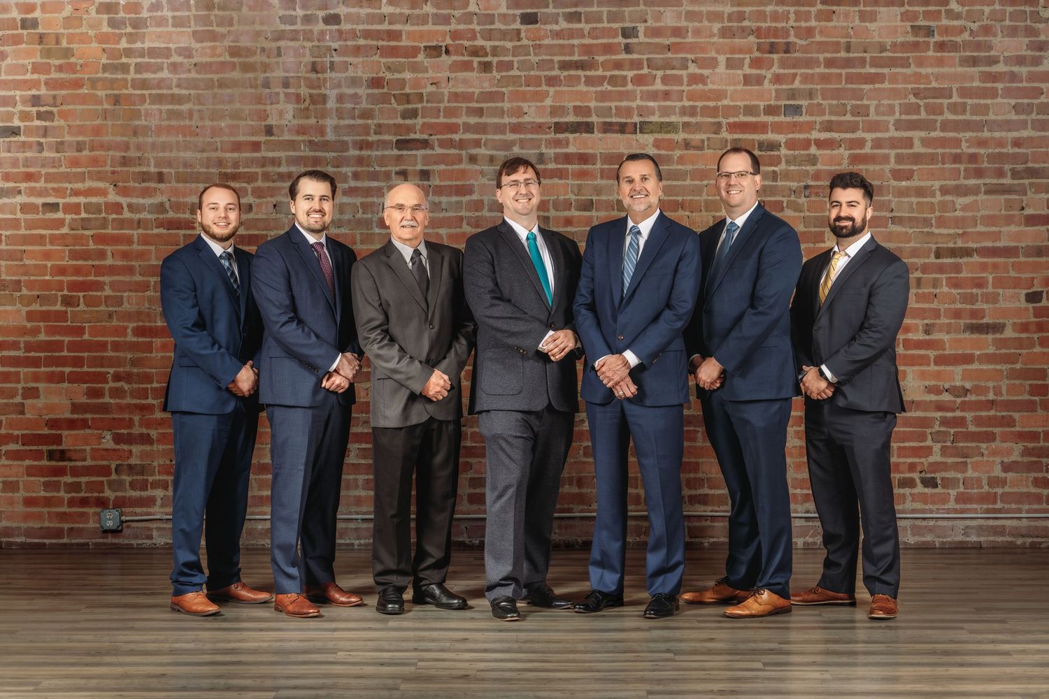 Seven professional men in suits pose for a formal group photo against an exposed brick wall backdrop.