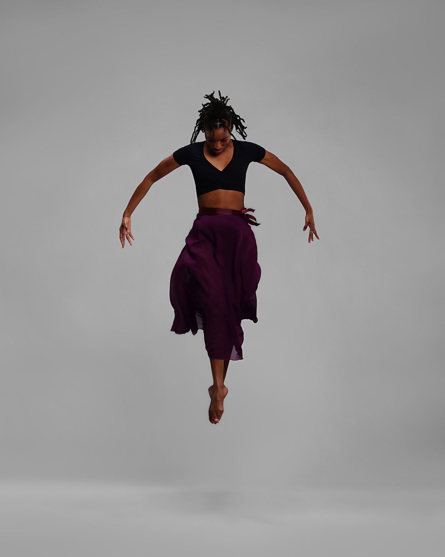 A dancer leaps gracefully in a black crop top and flowing purple skirt against a gray studio background.