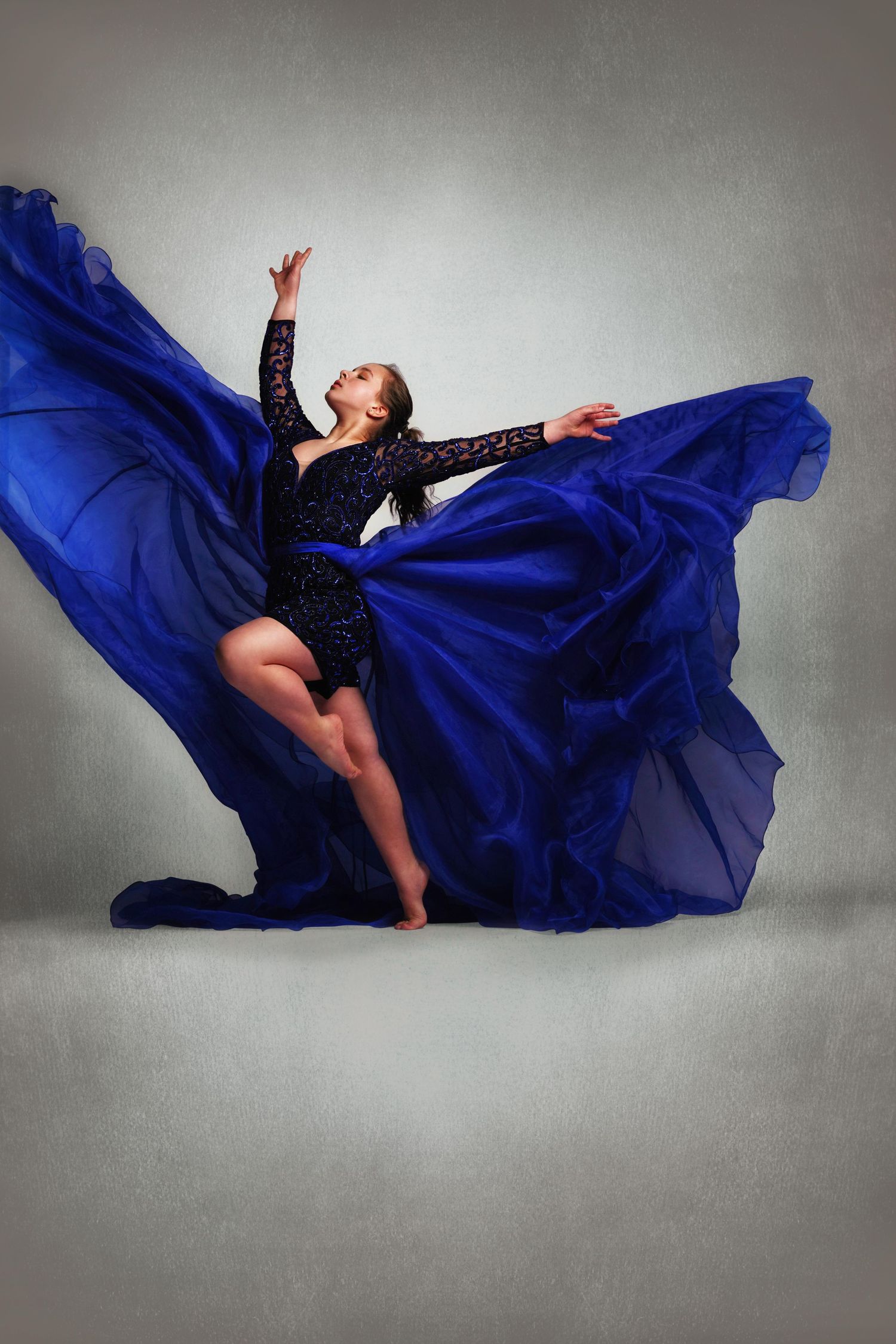 A dancer leaps gracefully in a flowing royal blue gown, fabric billowing dramatically against a soft gray studio background.