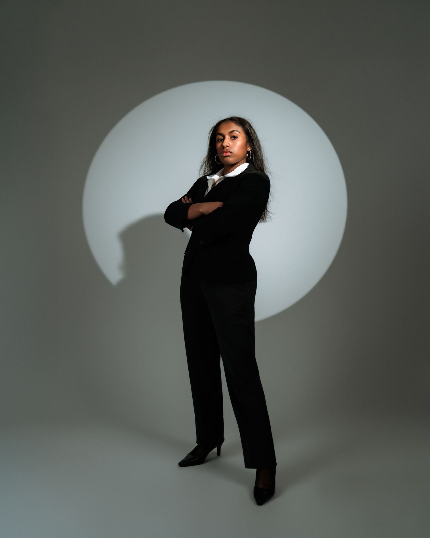 Woman in black suit with arms crossed stands confidently before a circular light backdrop.