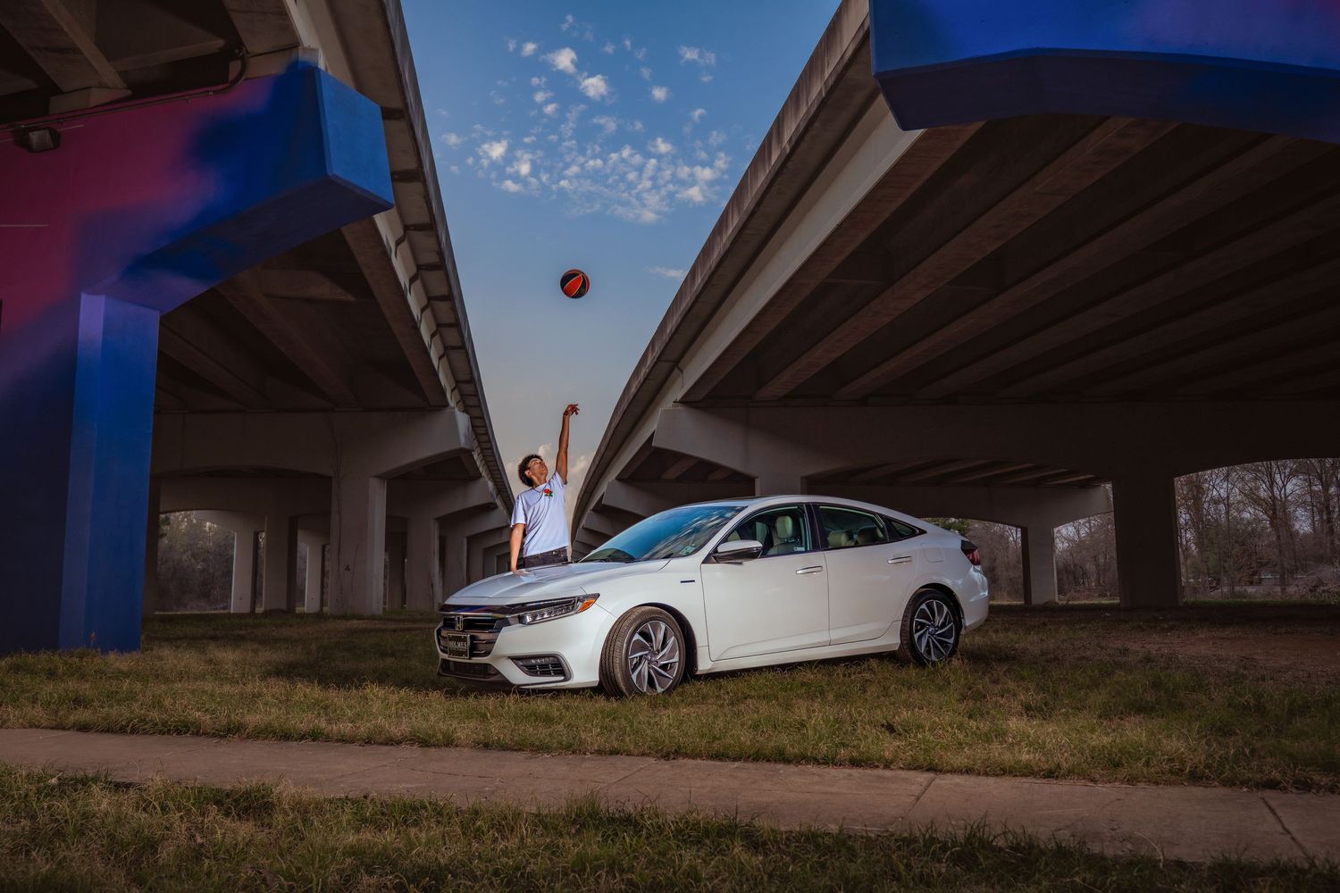 Person tossing a basketball beside a white Honda Insight sedan under highway overpasses at dusk.