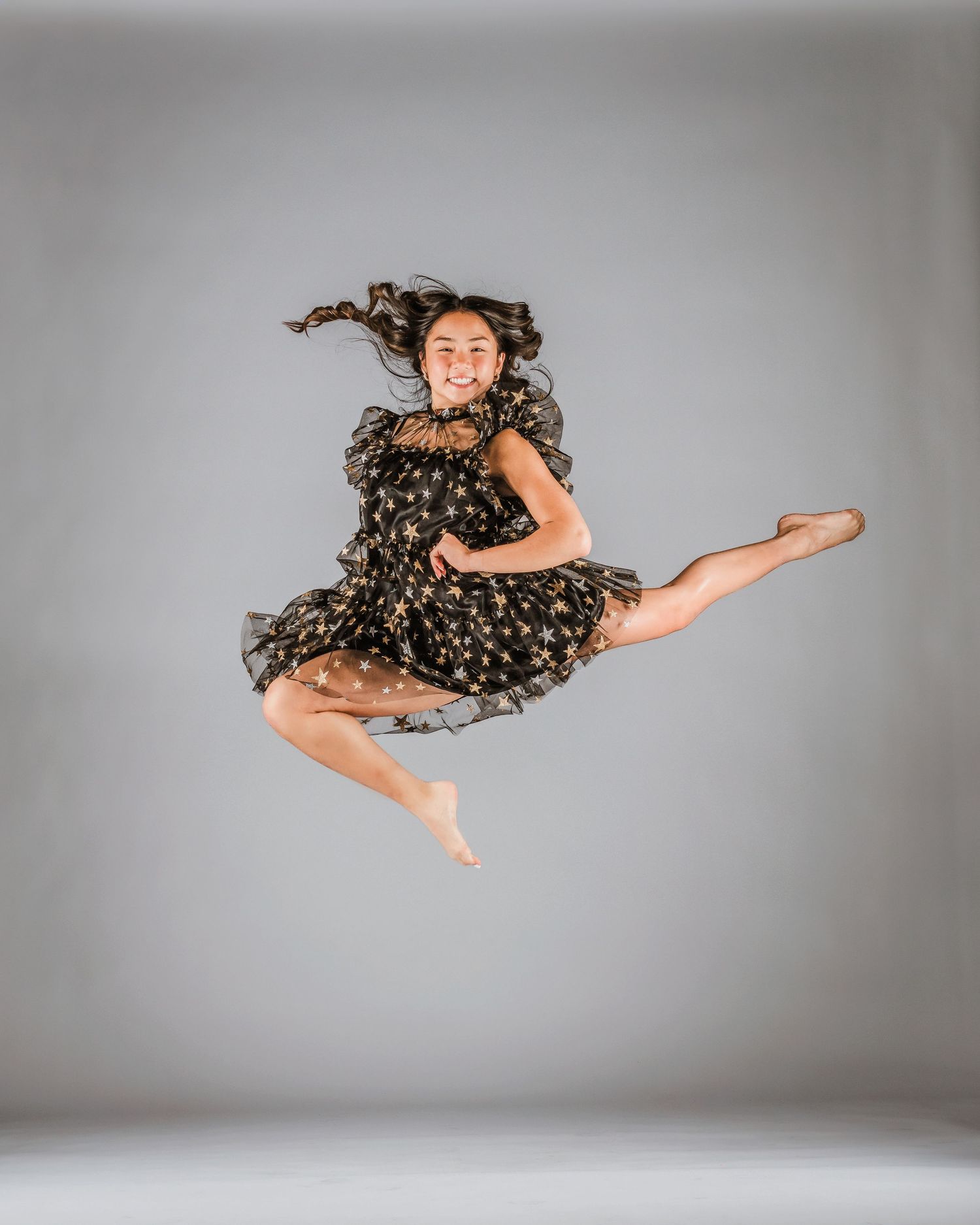 Young dancer leaps gracefully in black star-print dress, hair flowing, one leg extended, smiling against gray backdrop.