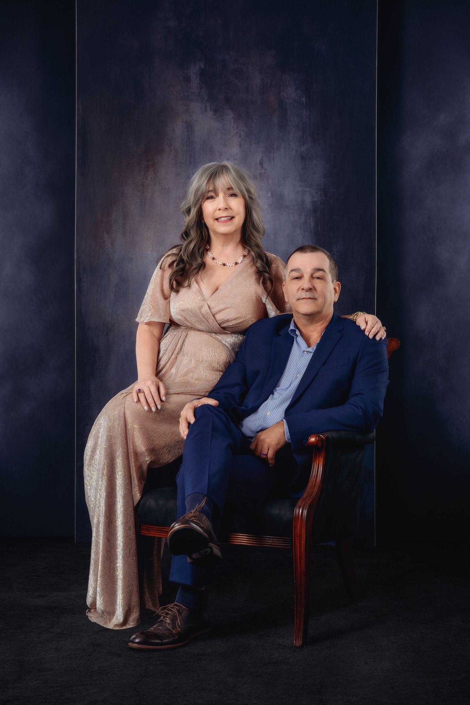 Elegant couple posing in studio, woman in beige gown standing beside seated man in navy suit, dark backdrop.