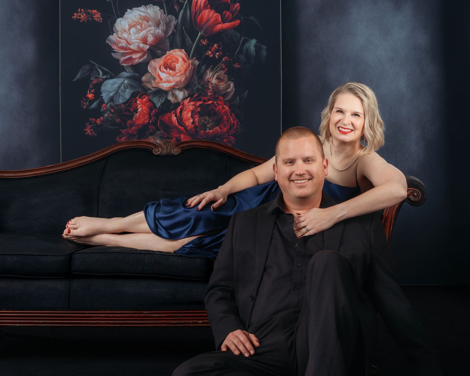 A smiling couple poses elegantly in front of a floral painting, woman in blue dress reclines on dark sofa, man in black sits below.