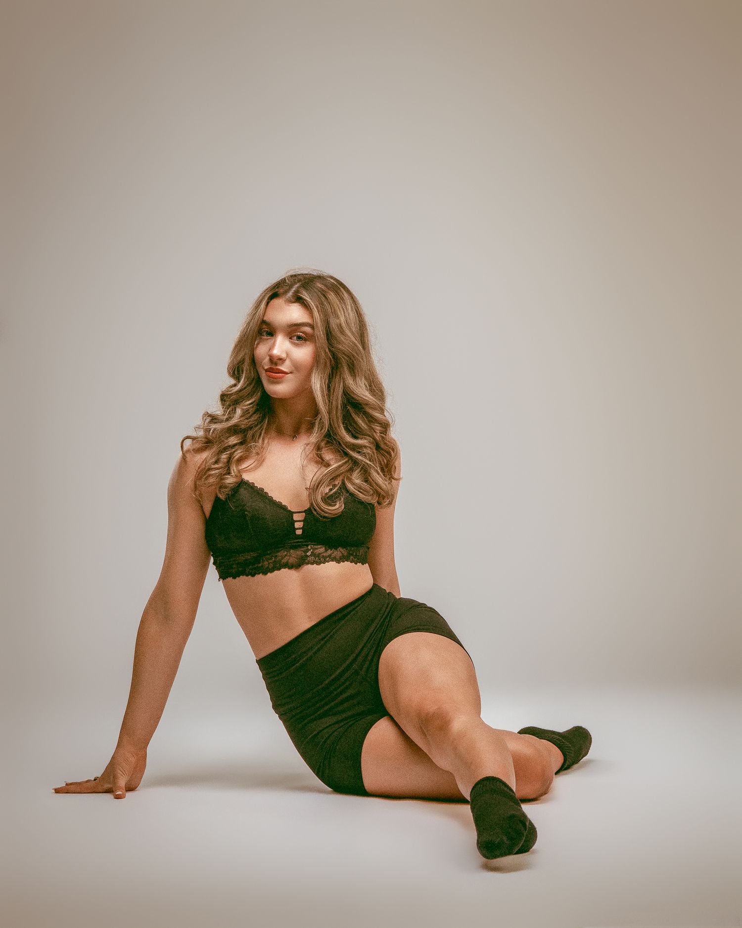 Young woman in dark green lace bralette and mini skirt posing seated on white studio floor.