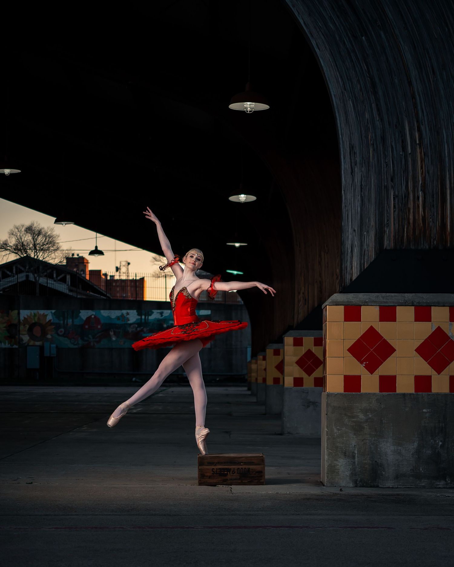 Ballerina in red tutu performs an arabesque on pointe in an urban underpass setting.