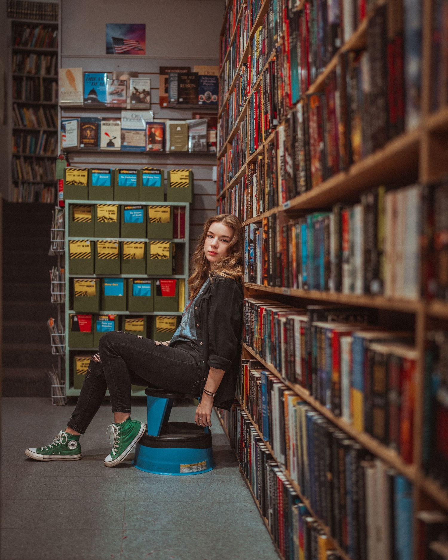 Young woman in black jeans and green Converse sitting on a stool between bookshelves in a bookstore.