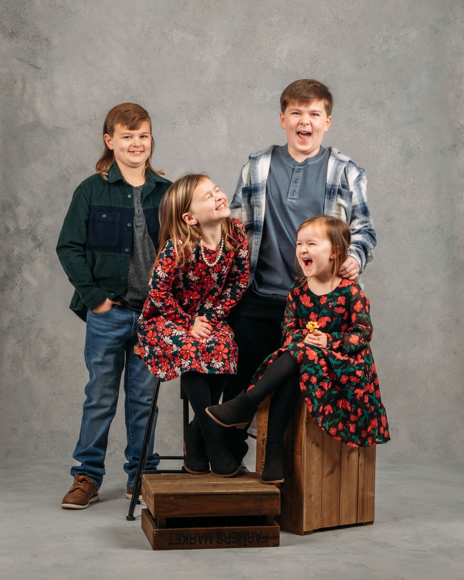 Four laughing children pose playfully on wooden crates in a studio portrait setting.