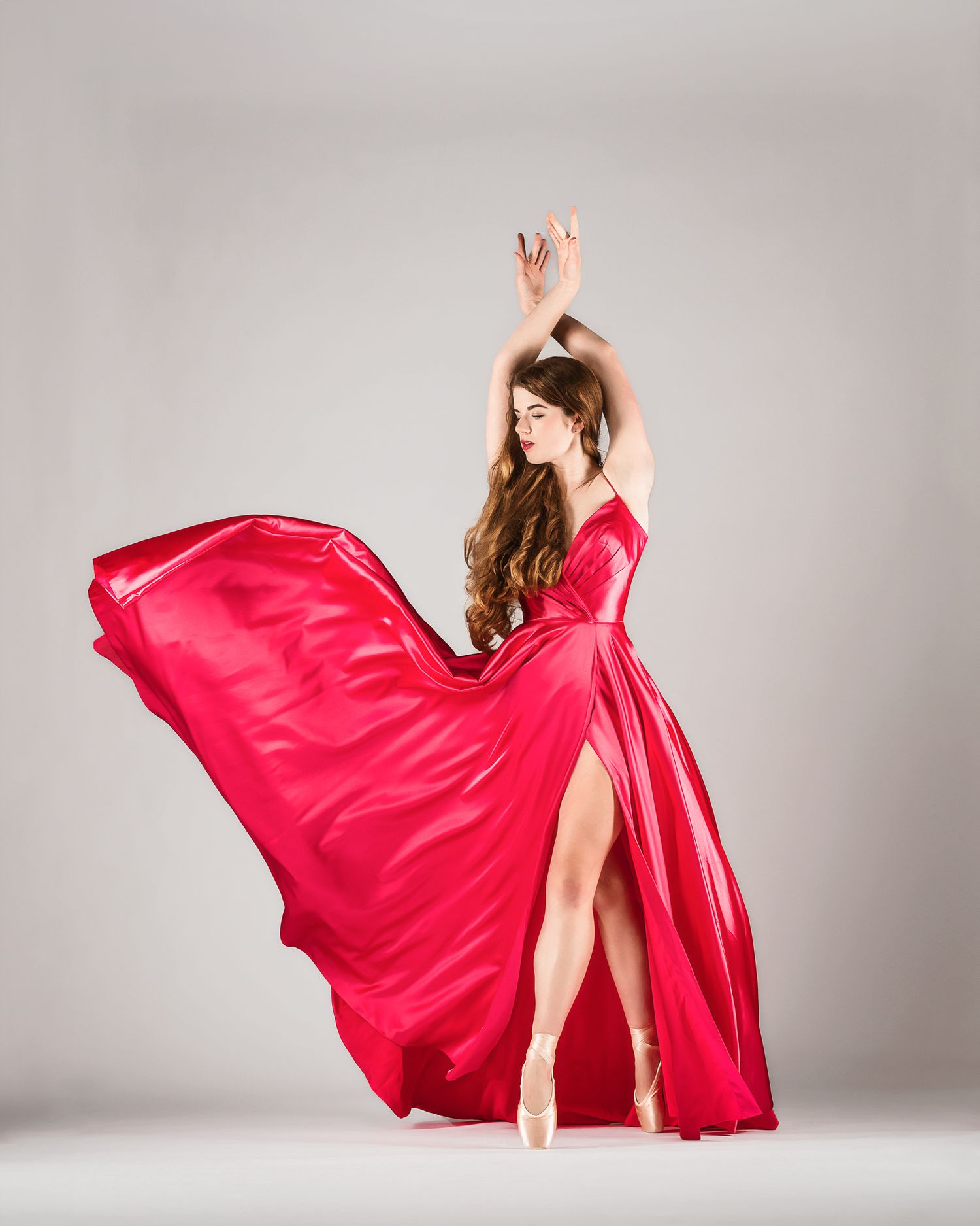 Ballerina in flowing red satin gown dances gracefully against white studio backdrop.