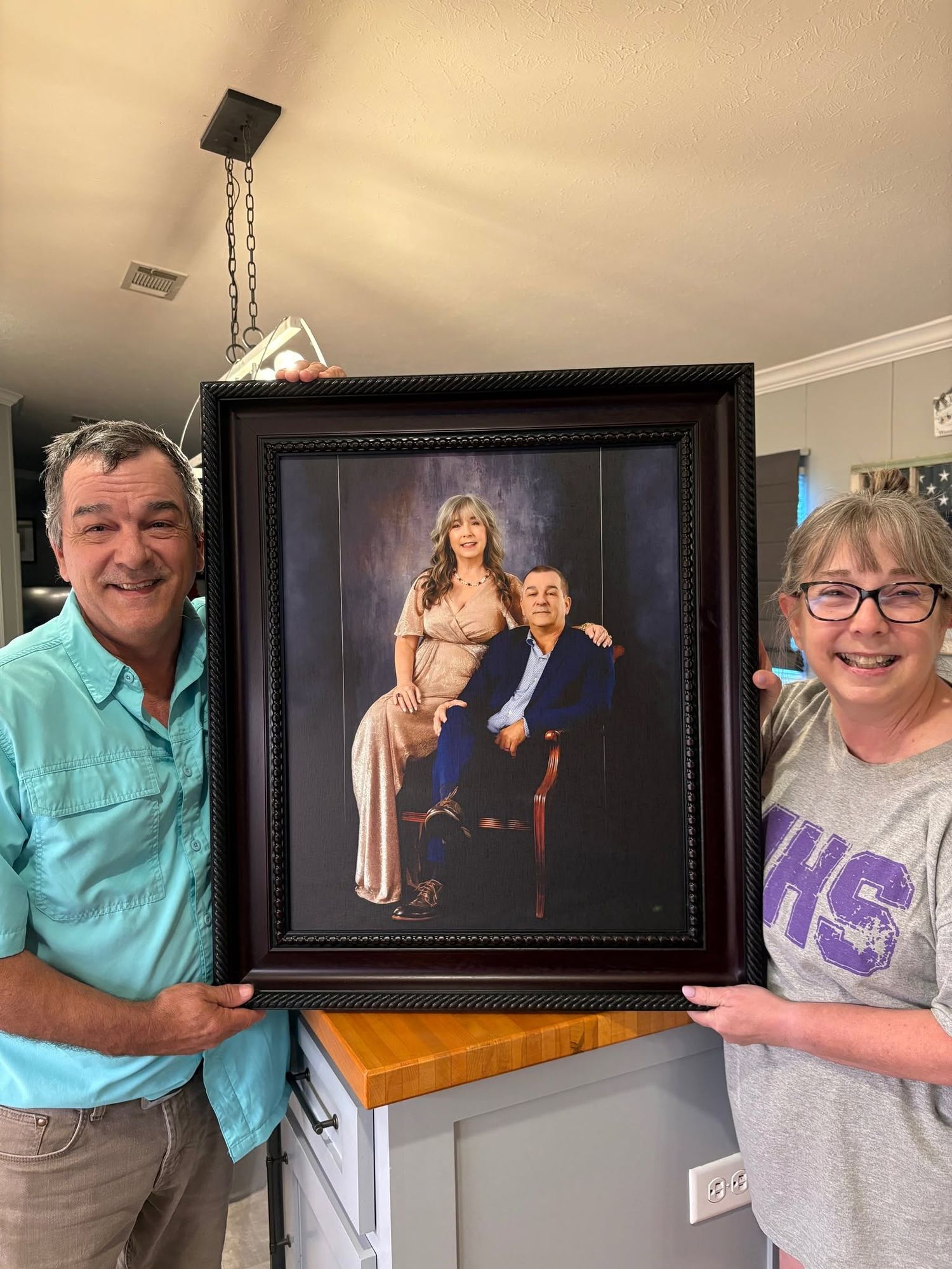 Couple proudly holds a large dark-framed formal portrait in their home kitchen.