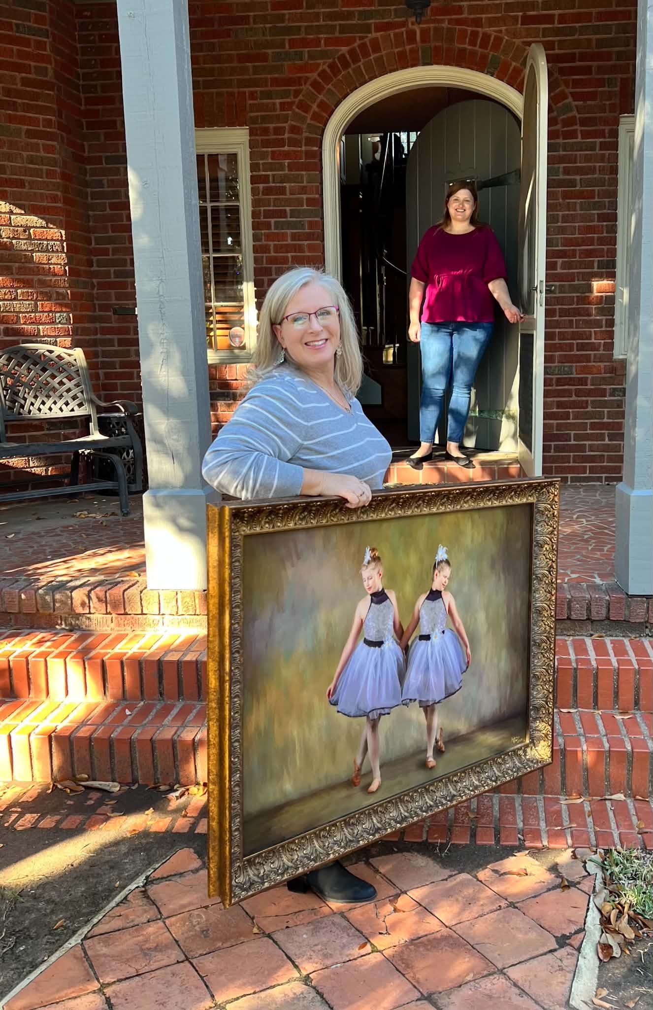 Woman holds a gold-framed painting of two young ballerinas in lavender tutus at a brick home.