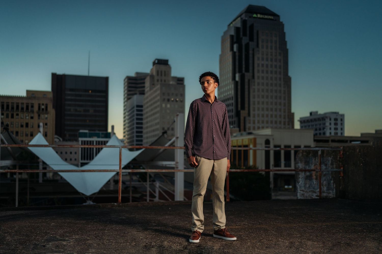 Young man standing on rooftop at dusk with city skyline buildings in background.