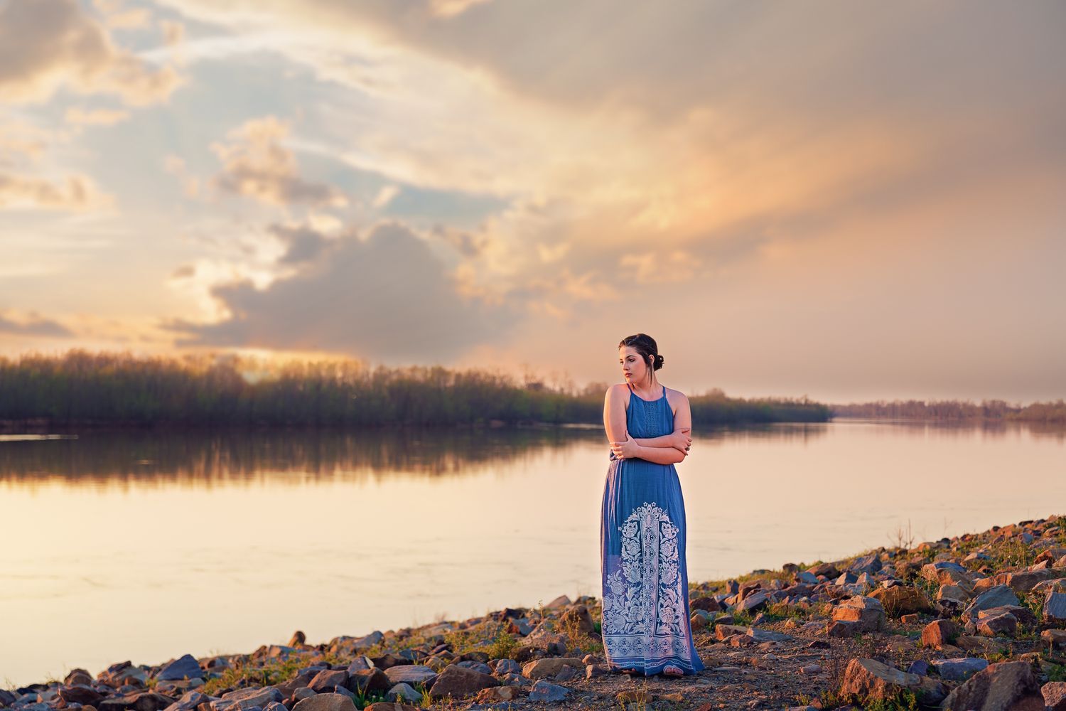 Woman in a blue maxi dress standing by a rocky lakeside during a stunning golden sunset.
