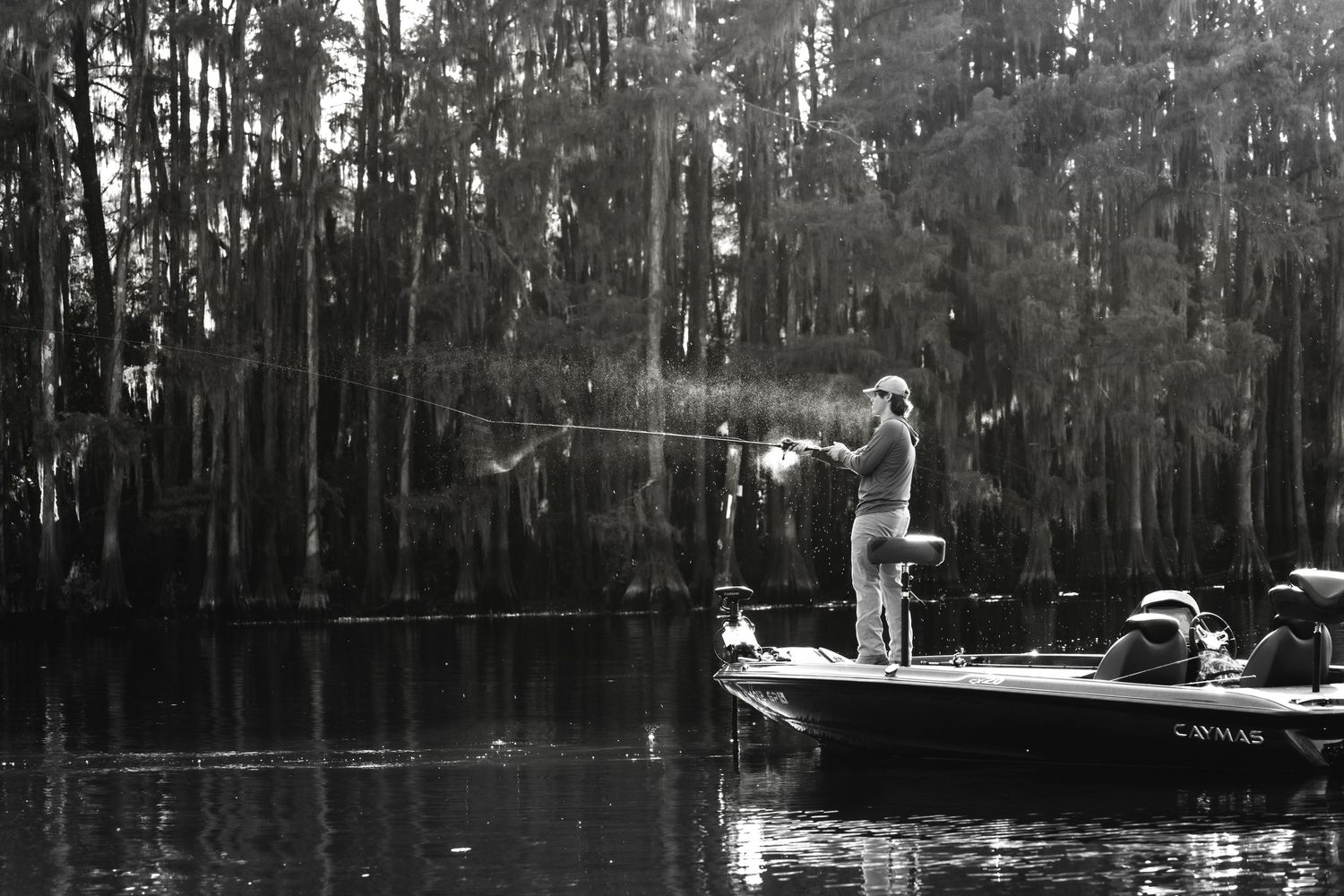 Black and white photo of a fisherman casting a line from a boat in a cypress swamp.