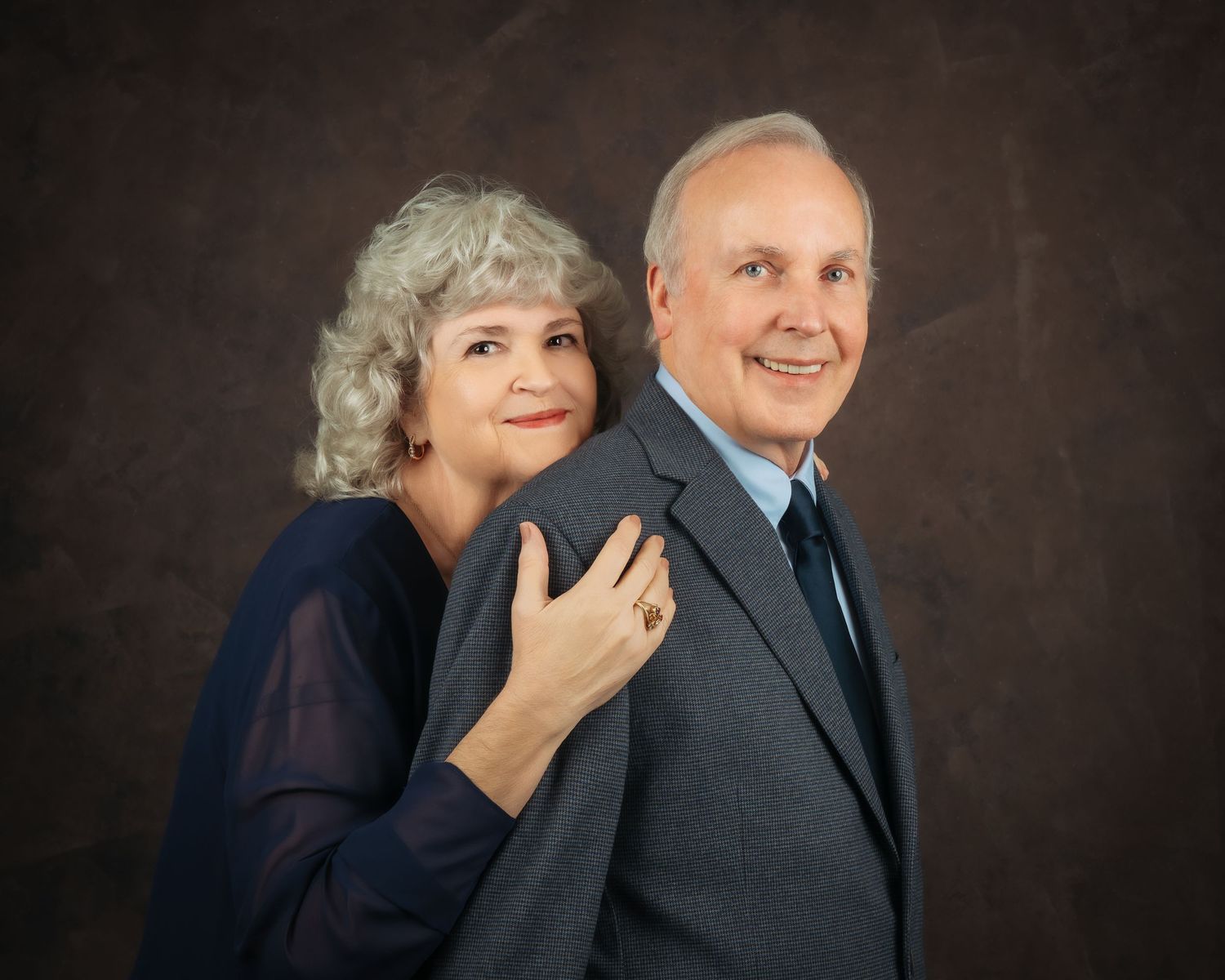 Older couple in a playful studio pose, woman in navy outfit resting hand on mans gray suit jacket.