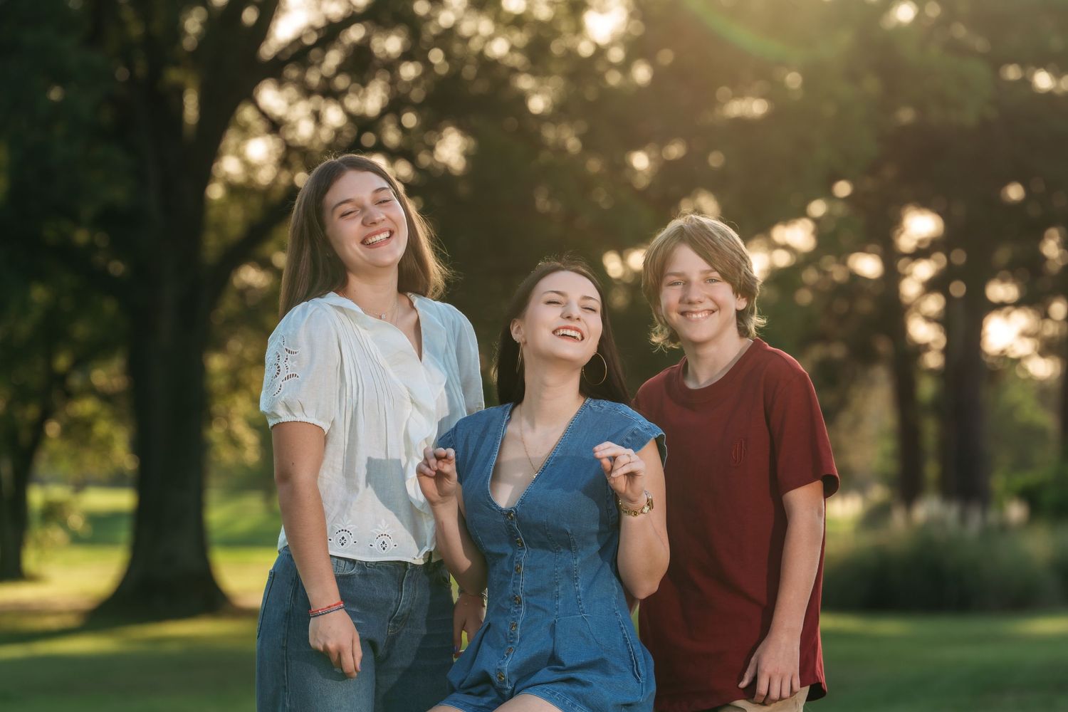 Three smiling teenagers laughing together outdoors in a sunny park with green trees behind them.