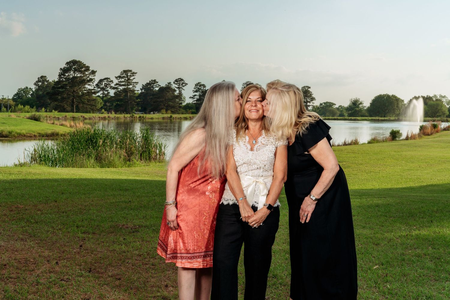 Three women sharing a warm embrace on a green lawn with a pond and fountain behind them.