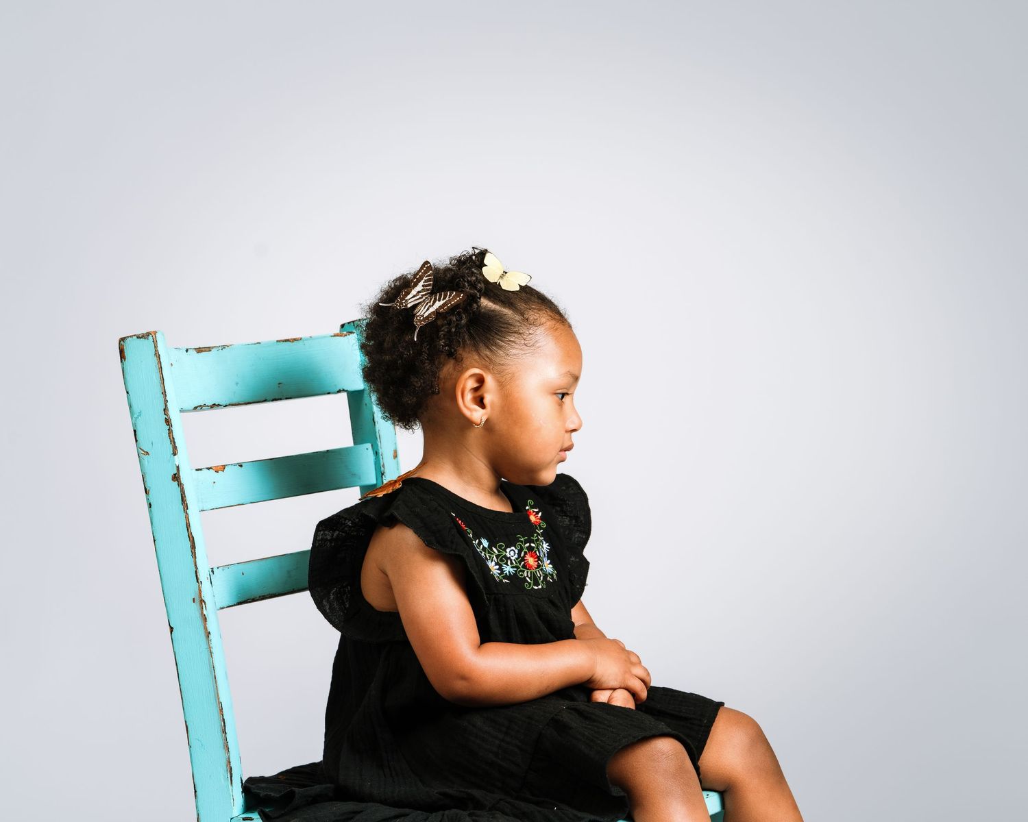 Young Black girl in black dress sits on teal chair, looking away, with hair accessory, against gray background.