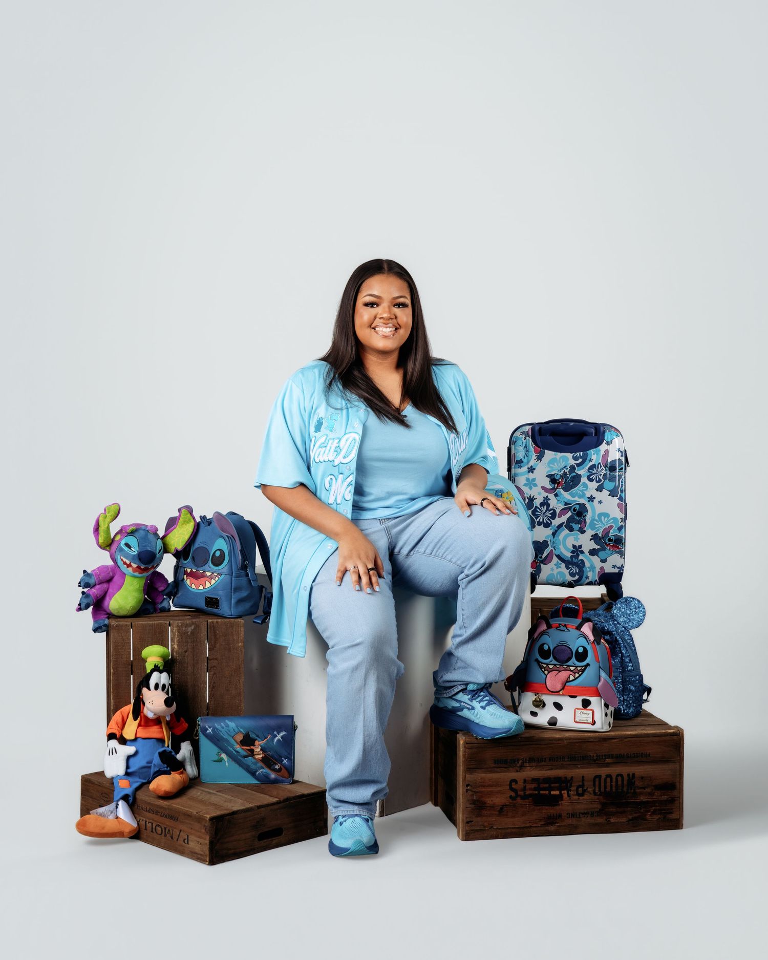 Woman in blue scrubs poses with Stitch-themed Disney merchandise, bags, and a Goofy plush on wooden crates.