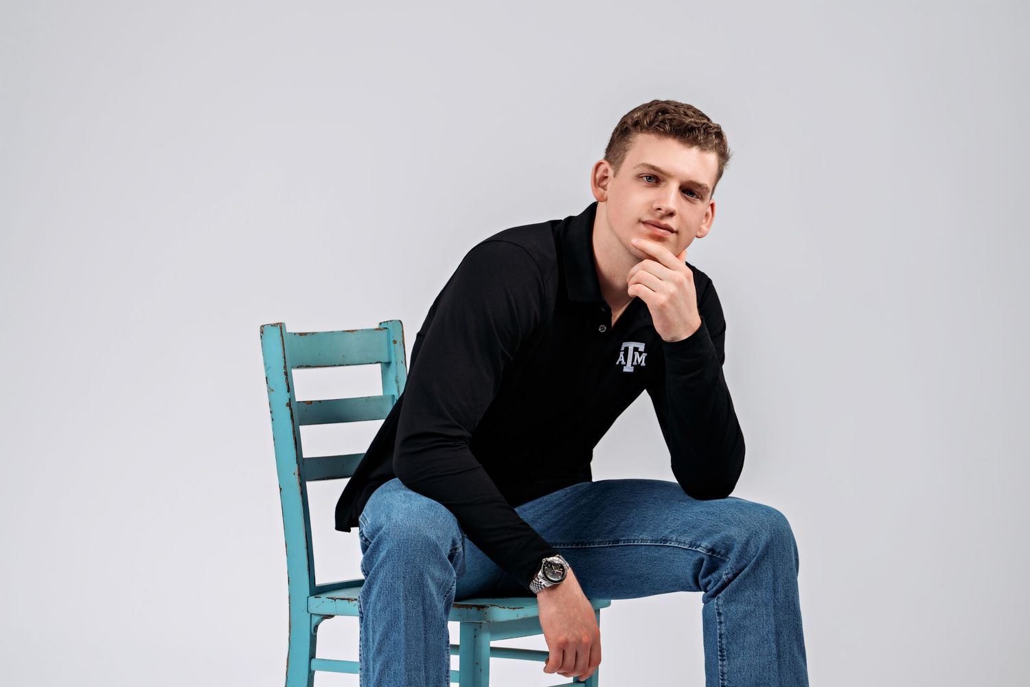 Young man in black Texas A&M polo sits thoughtfully on teal chair against white background.