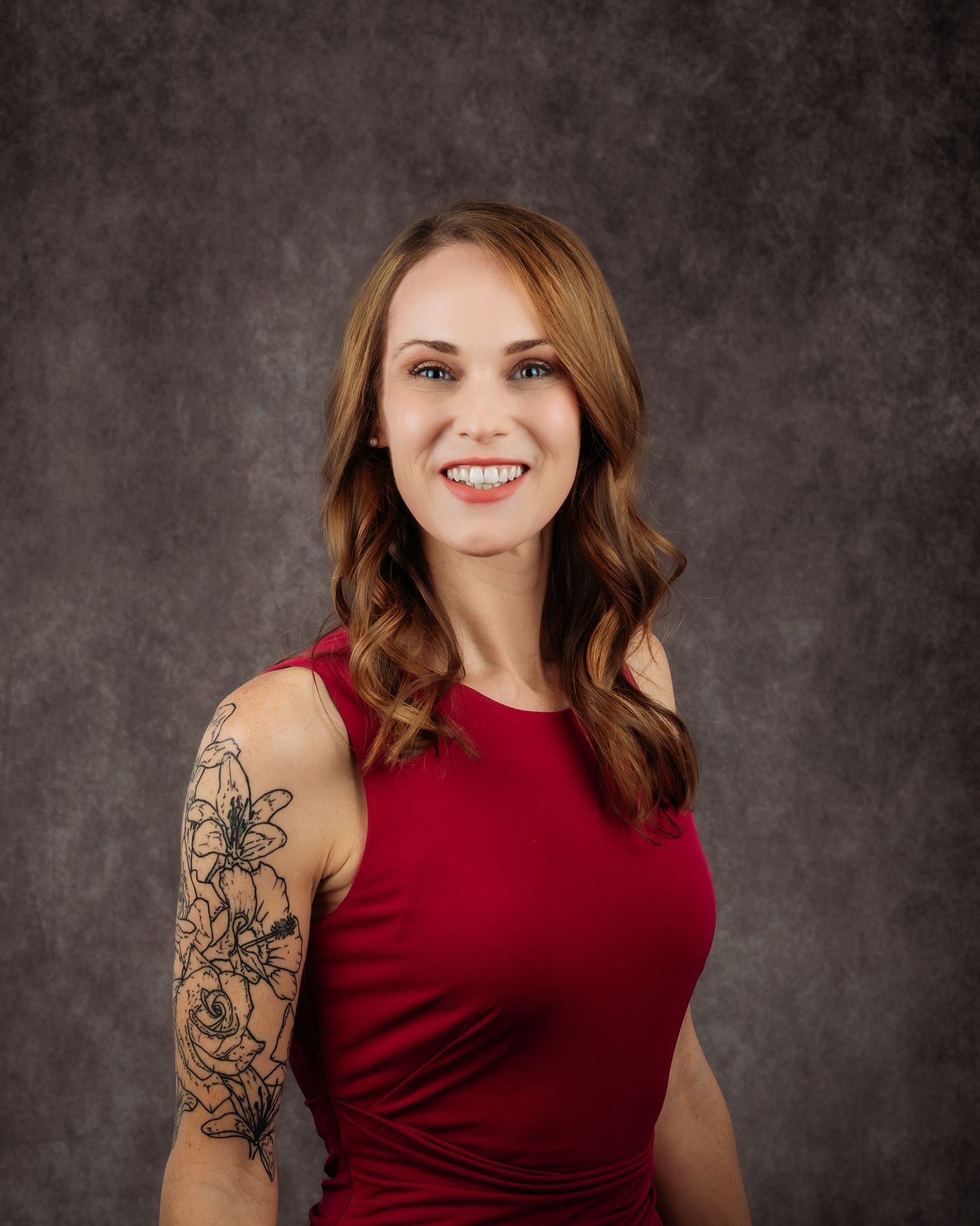 Professional headshot of a smiling woman in a red dress with floral arm tattoo against a gray backdrop.