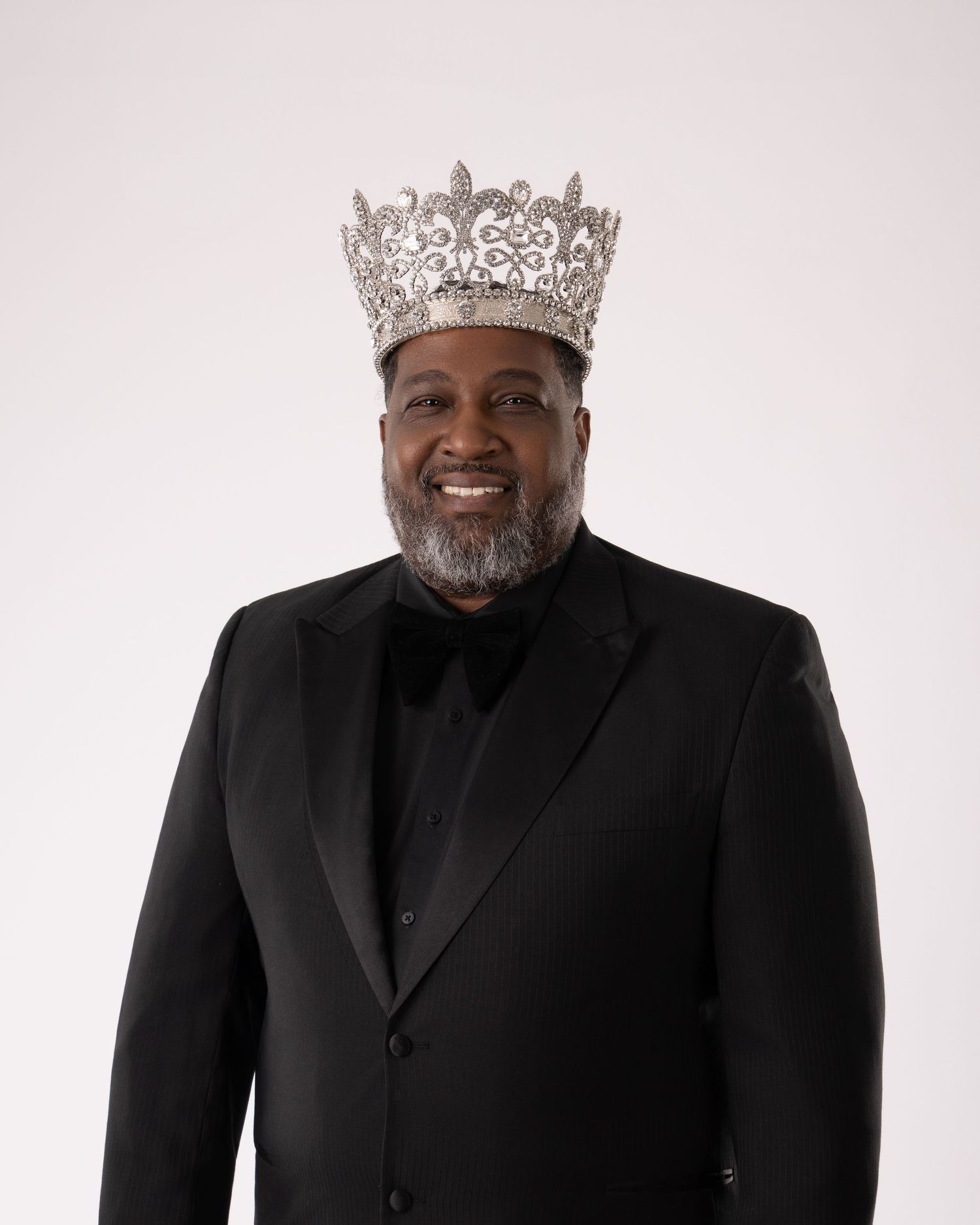 Man in black tuxedo wearing ornate silver crown smiling against white background.