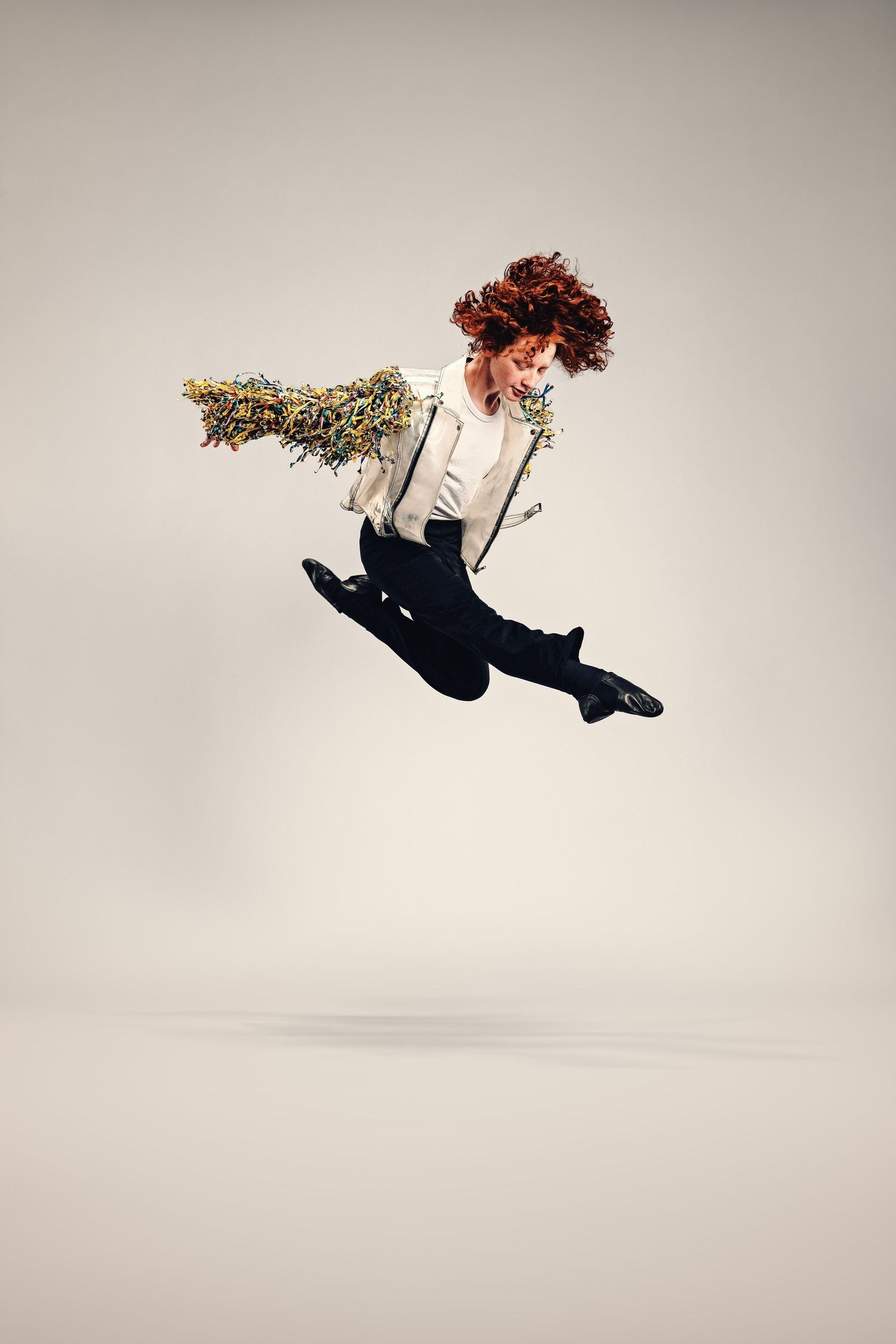 Redhead dancer mid-air jump wearing white shirt, suspenders, black pants, and floral accessories against gray background.