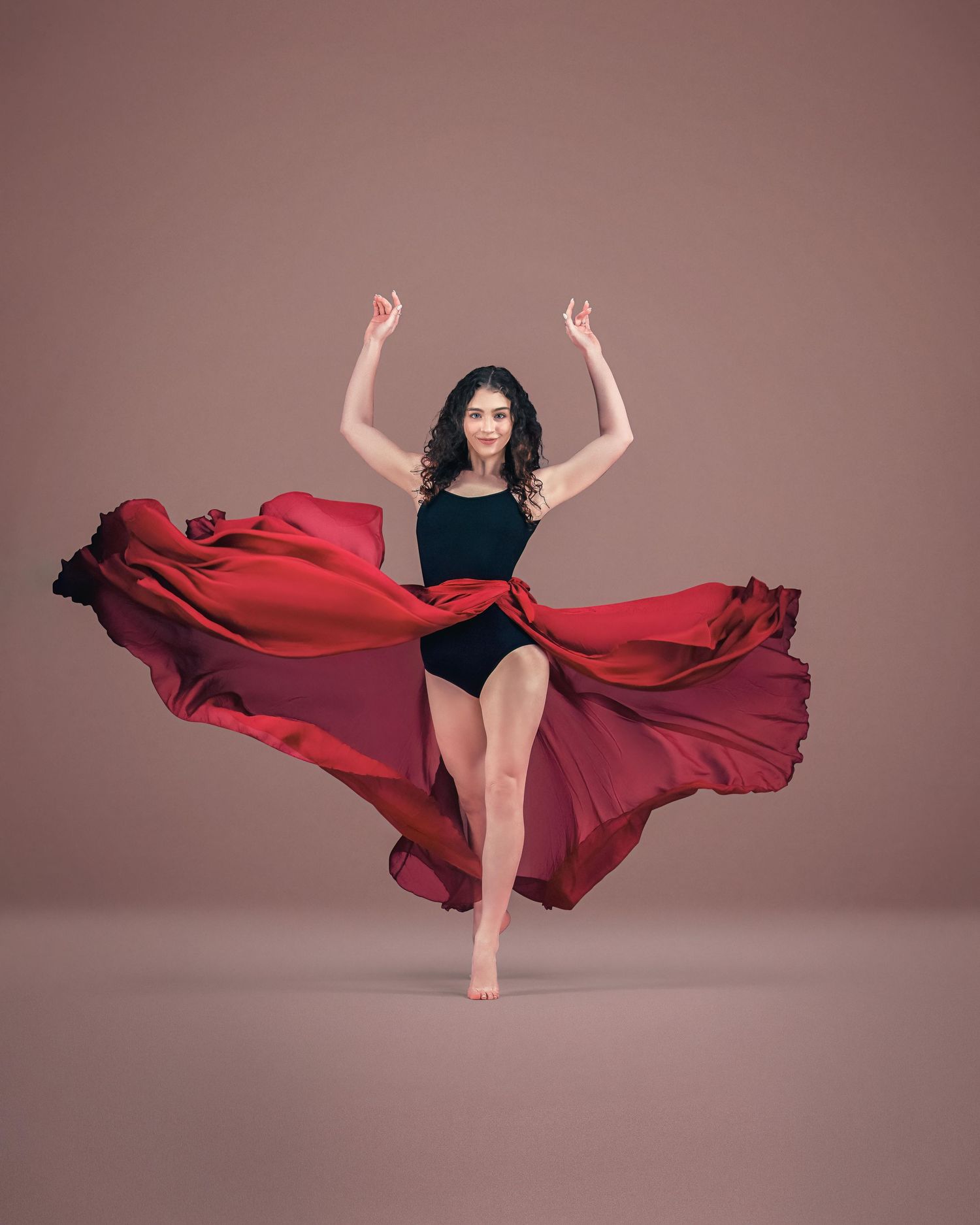 A dancer in a black leotard poses gracefully with a flowing red fabric billowing behind her on a mauve background.