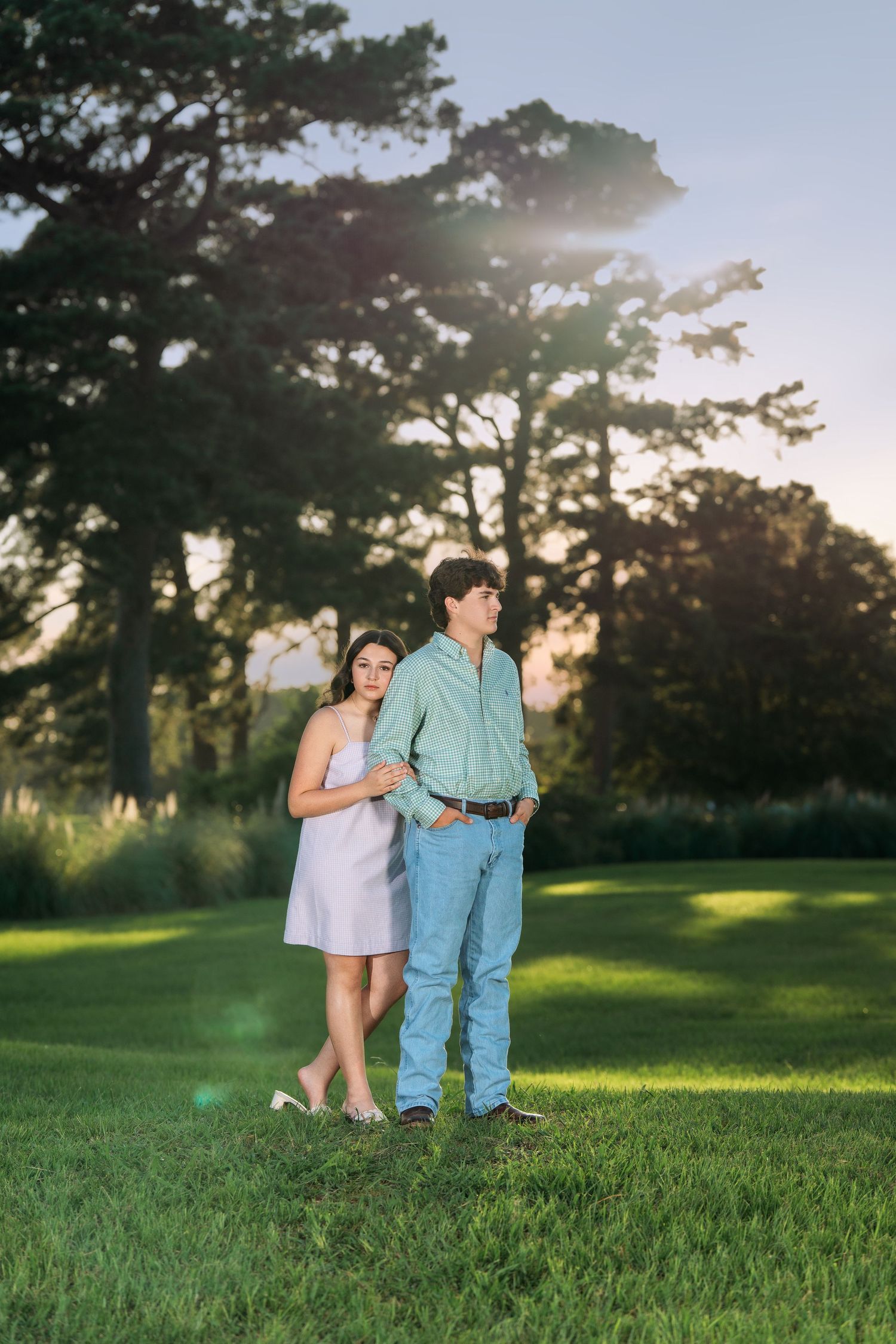 A couple stands together on a lush green lawn at golden hour with sunlit trees in the background.